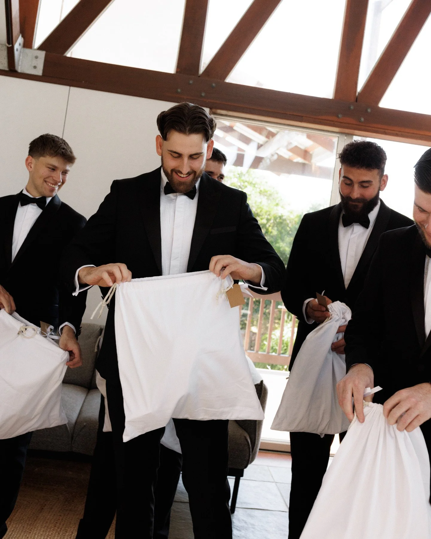Group of men dressed in tuxedos and bow ties opening white cloth bags inside a room with a high, angled ceiling and large windows, with greenery visible outside.