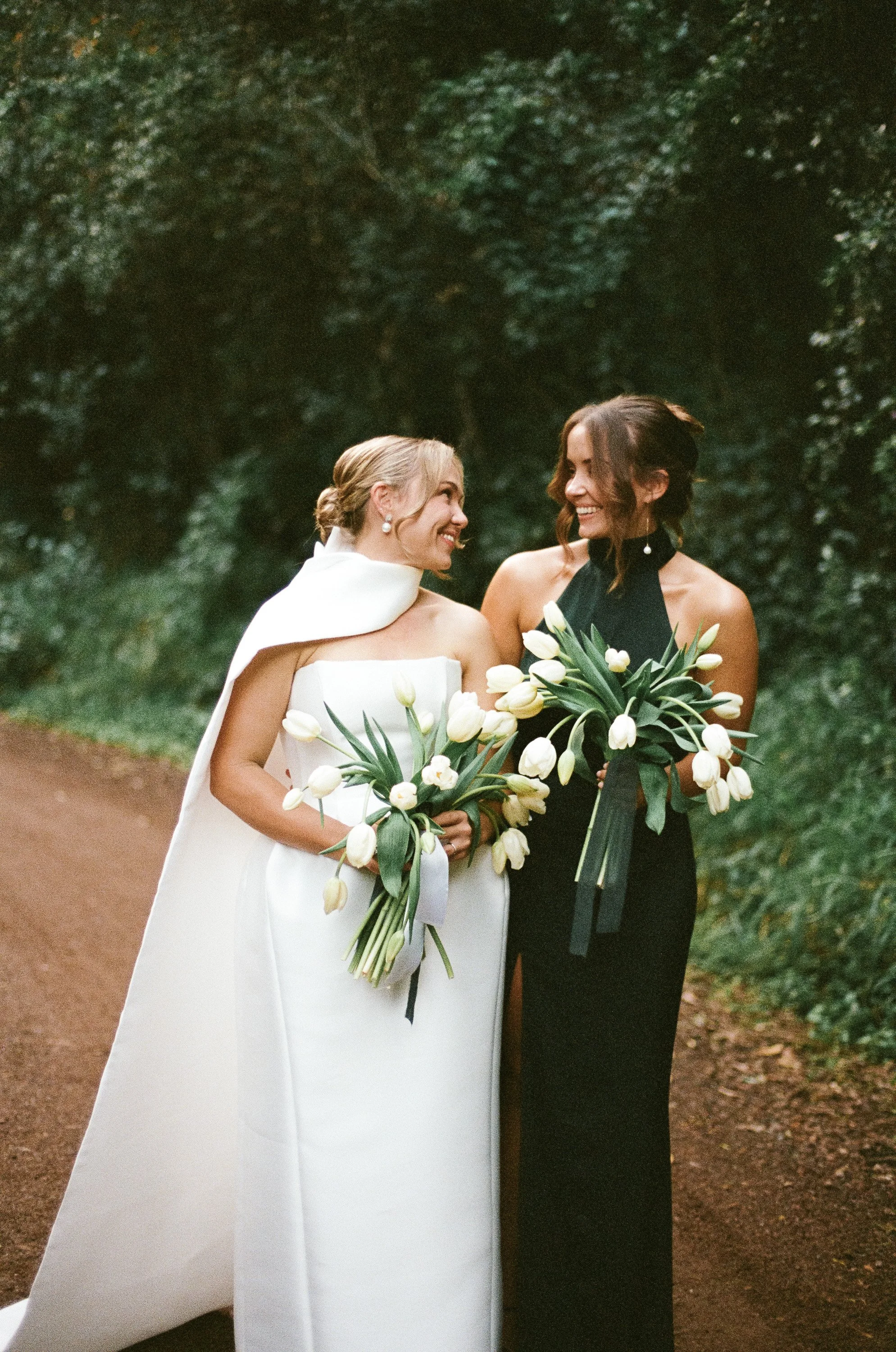 Two women standing outdoors, smiling at each other. One is wearing a white dress with a cape, and the other is in a black dress. Both hold bouquets of white tulips - Elevated Weddings shot on 35mm film