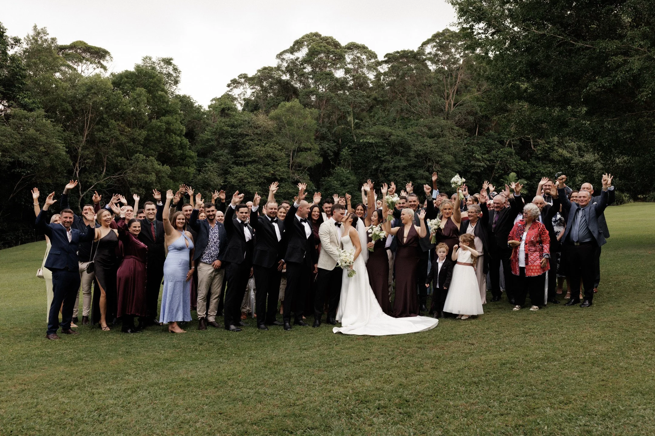 Group of wedding guests and couple posing outdoors on a grassy field with trees in the background.