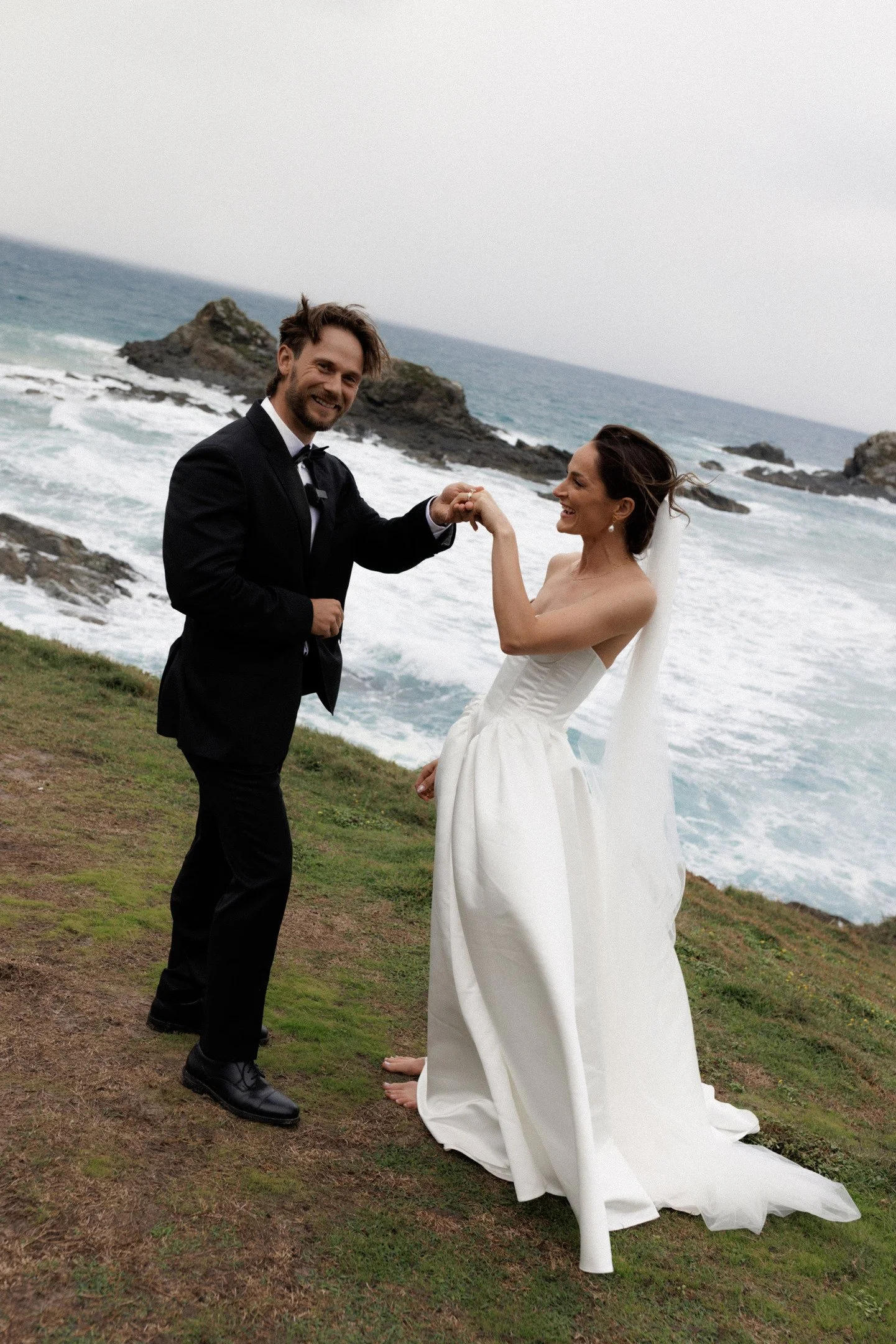 A bride and groom holding hands on a grassy area near the ocean, with rocks and waves in the background during a cloudy day.