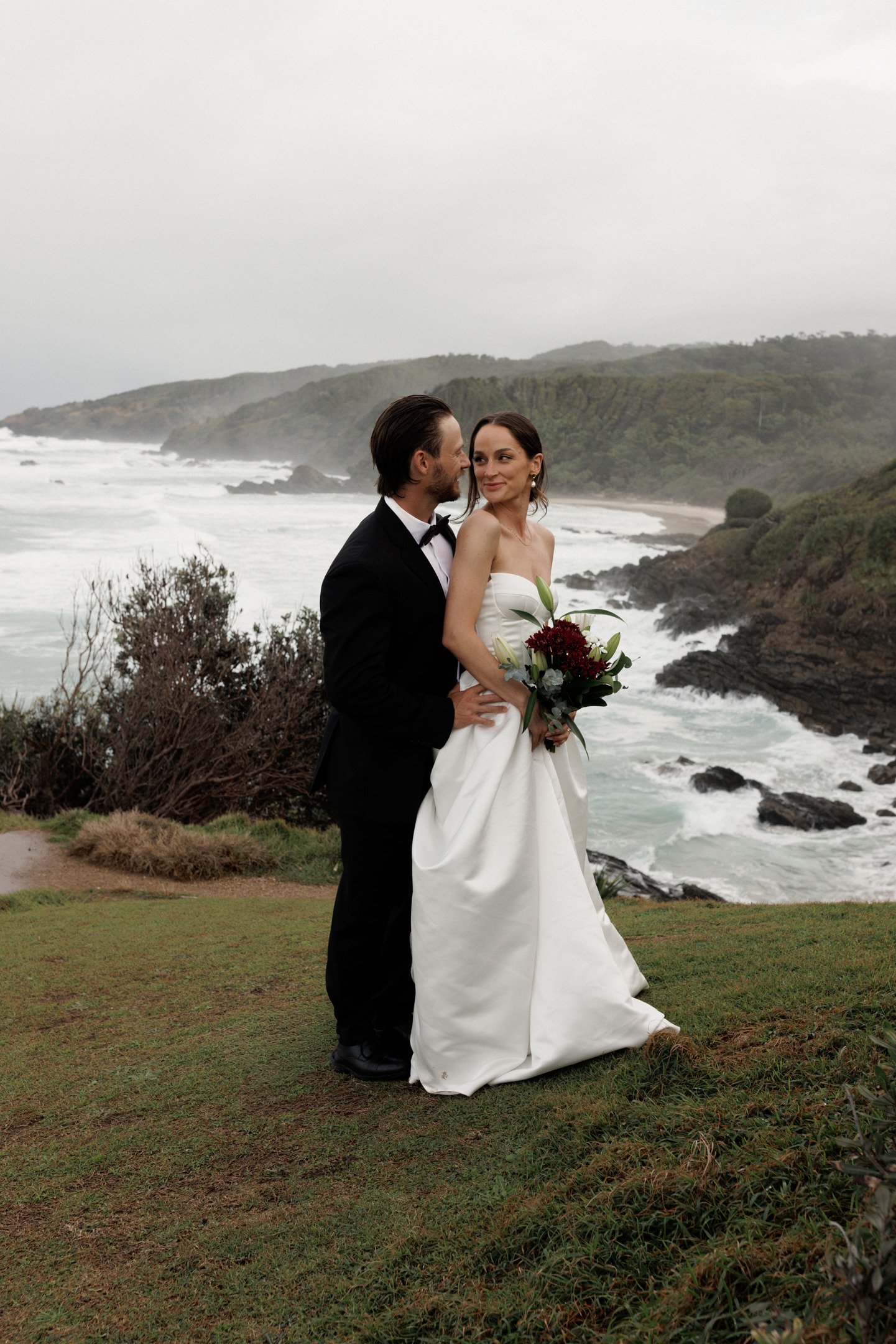 A bride and groom stand on a grassy area near the coast, with the groom in a tuxedo and the bride in a strapless white wedding dress holding a bouquet. They are looking at each other, with waves crashing on rocks and a hilly landscape in the background on a cloudy day.