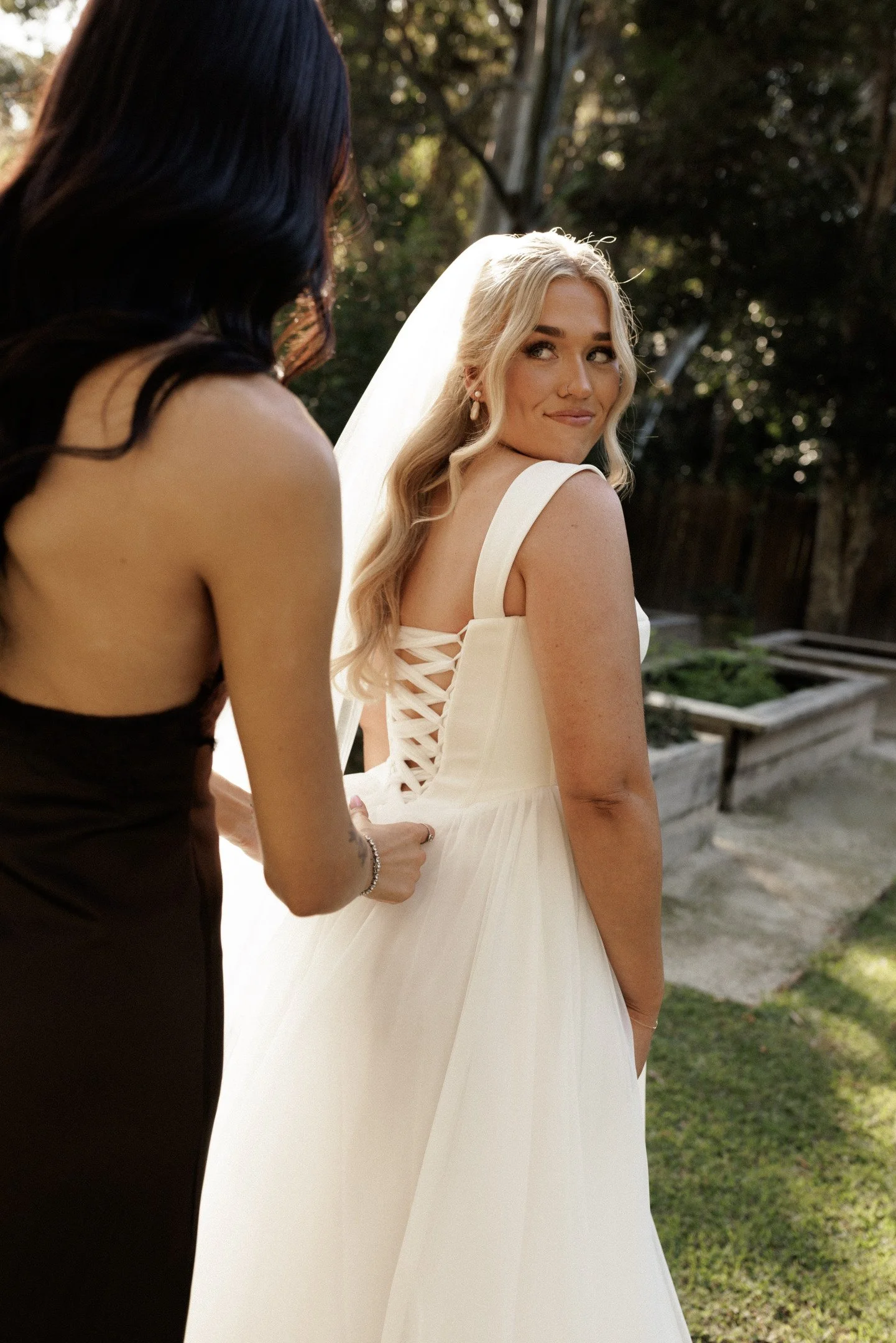 A bride with long blonde hair in a white dress standing outdoors with trees in the background, smiling while someone adjusts her dress.