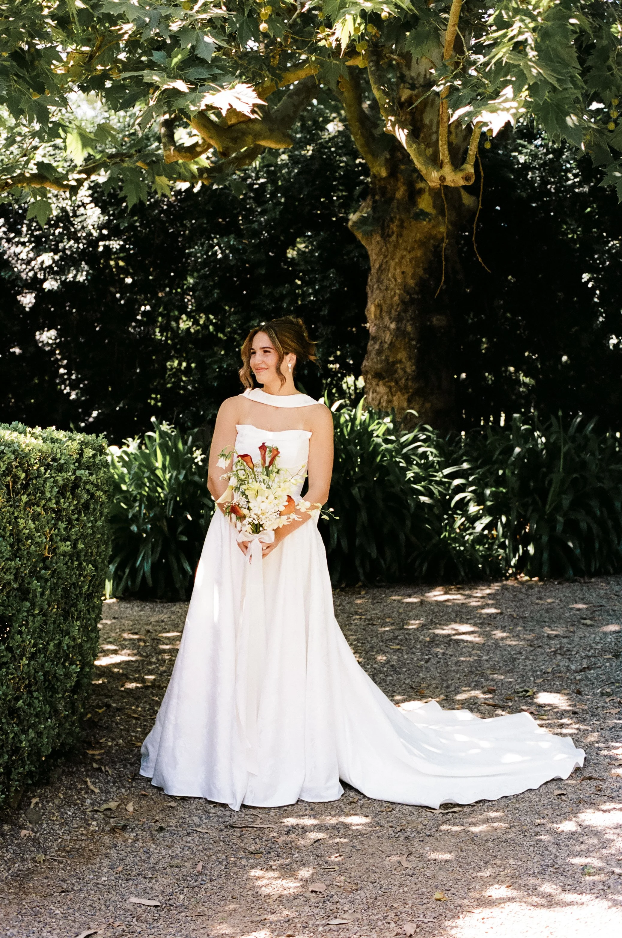 A bride in a white wedding dress holding a bouquet, standing outdoors in front of lush green foliage and a large tree.