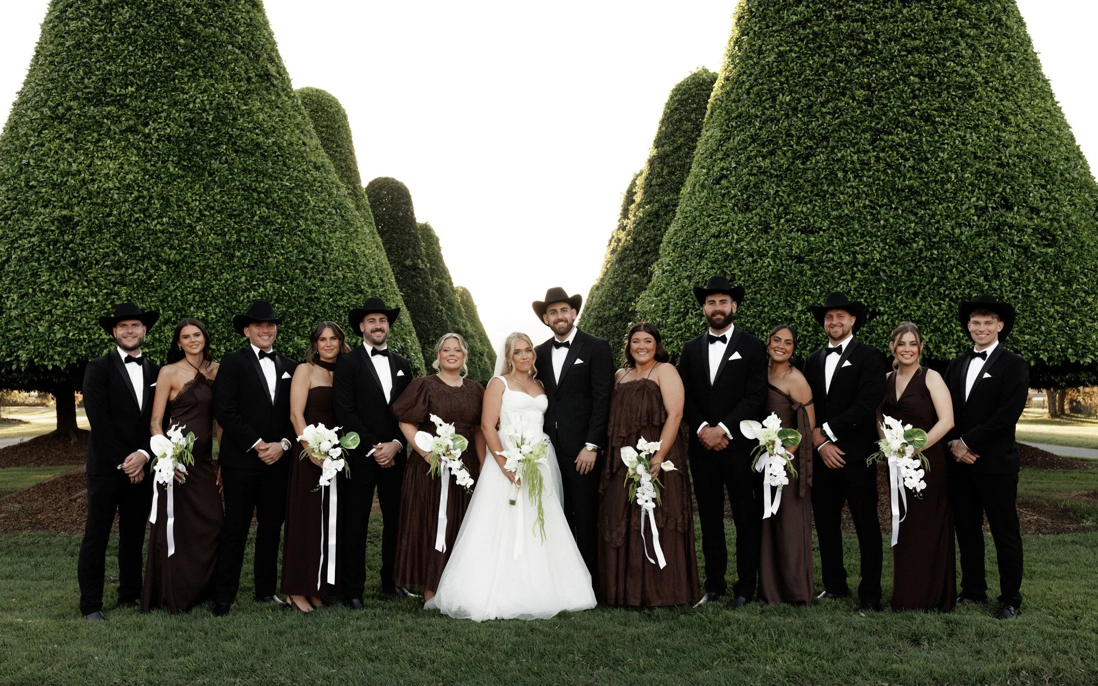 Group of wedding party members dressed in formal attire with some men in black tuxedos and some women in brown dresses, standing outdoors in front of large, well-trimmed green trees, with a bride in a white wedding dress and a groom in a black suit a