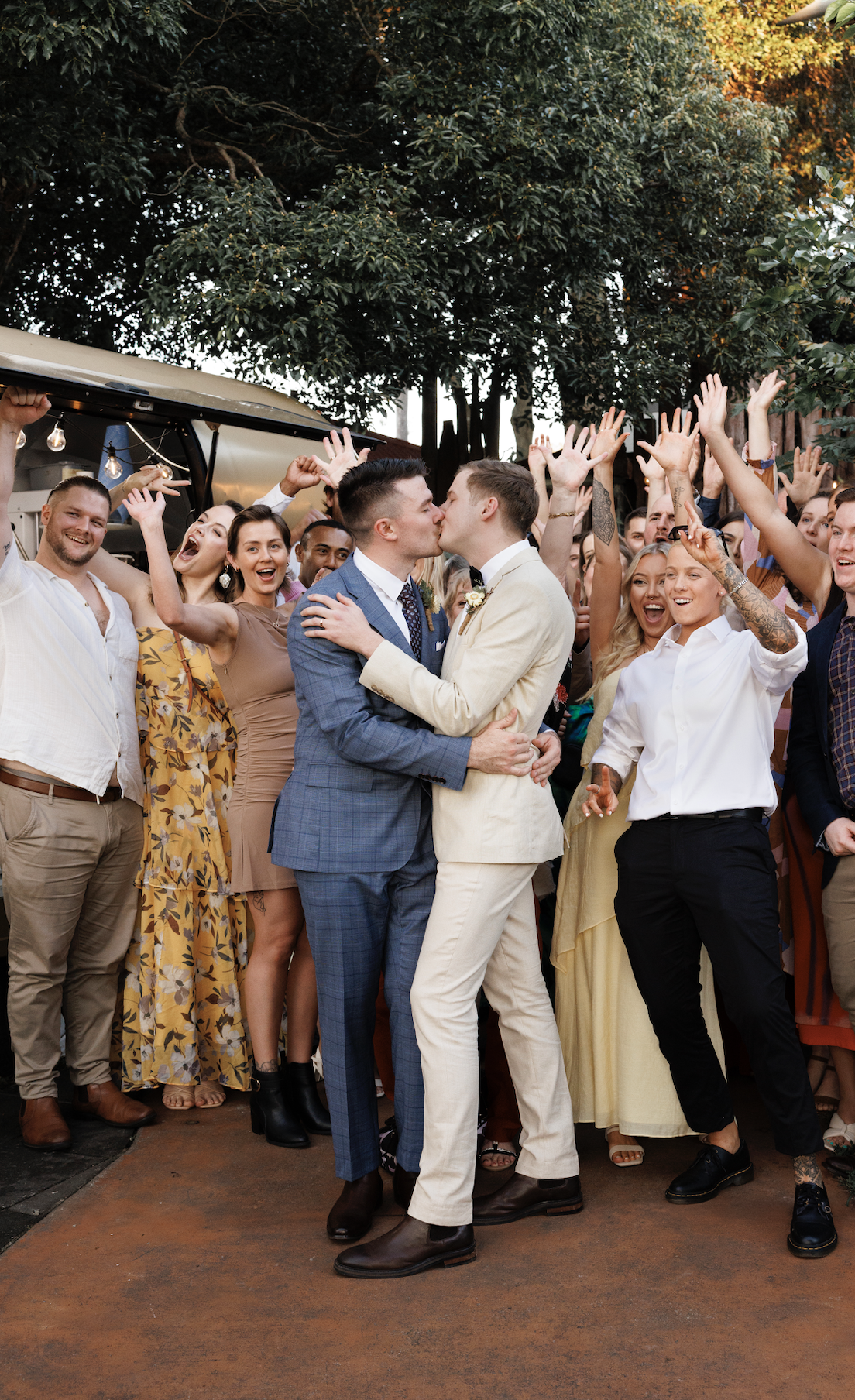 Two men in suits kissing at a celebration with a group of cheering, smiling people around them.