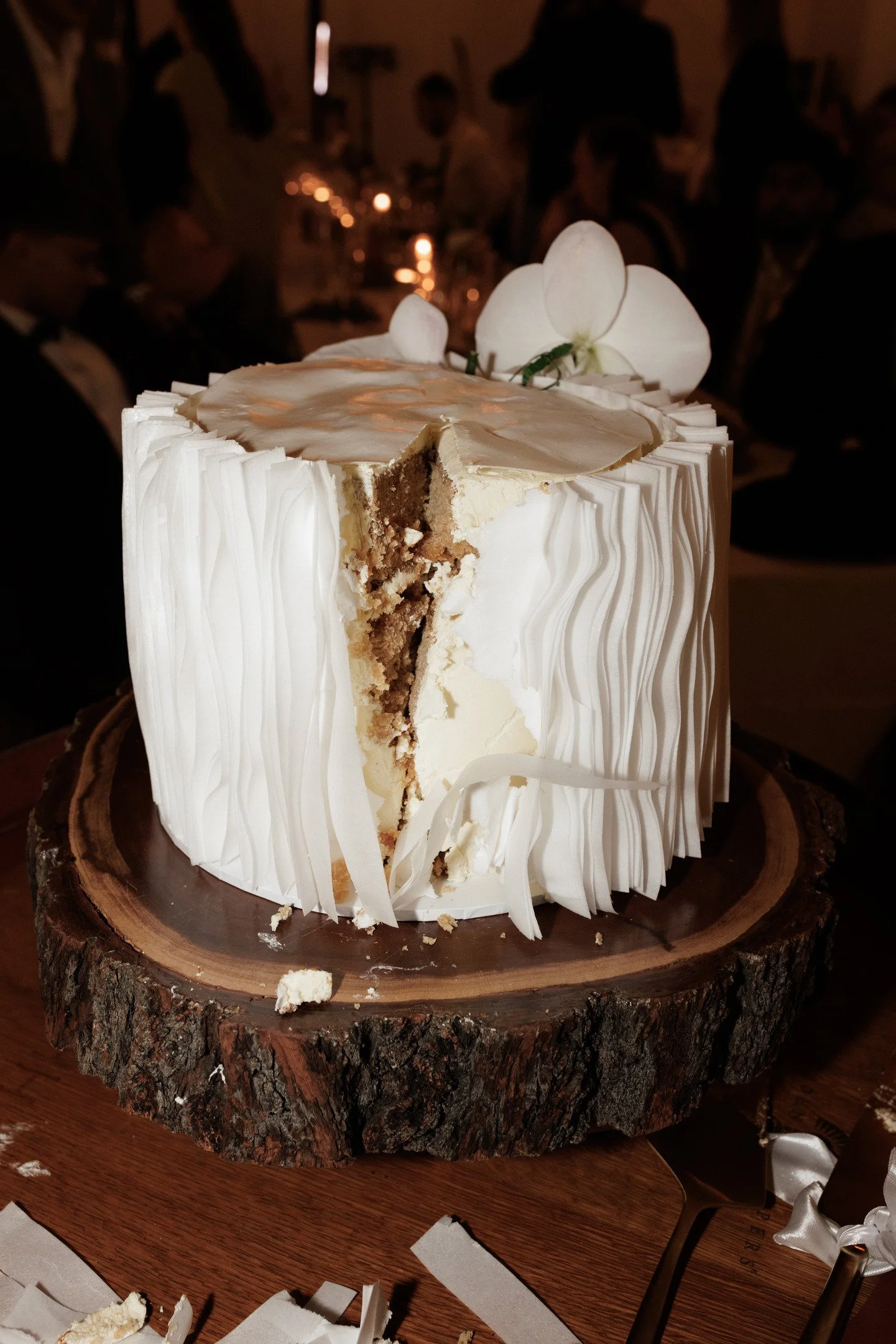 A partially cut white cake with a textured vertical stripe pattern on the sides, topped with white orchids, placed on a round wooden slab with a dark table beneath, and a blurred background with people at a gathering.