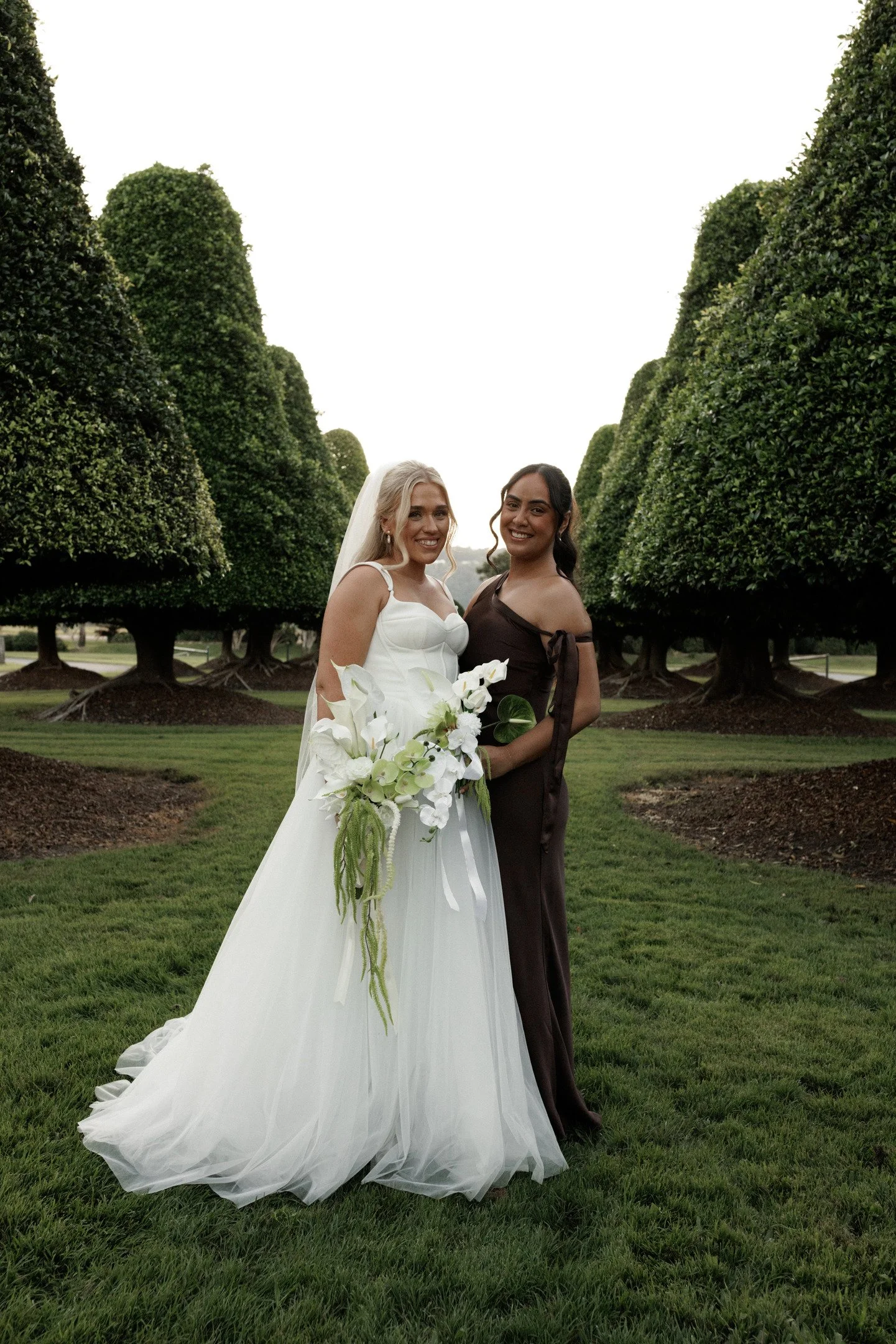 Two women standing in front of neatly trimmed green trees, smiling at the camera, one in a white gown holding a bouquet, the other in a dark dress.