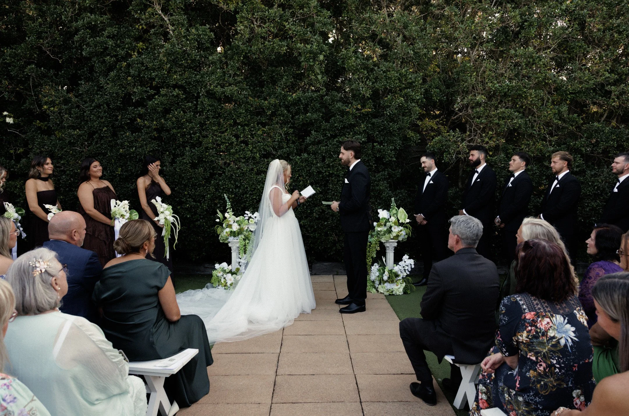 A bride and groom exchanging vows during an outdoor wedding ceremony, surrounded by seated guests and a lush green hedge backdrop.