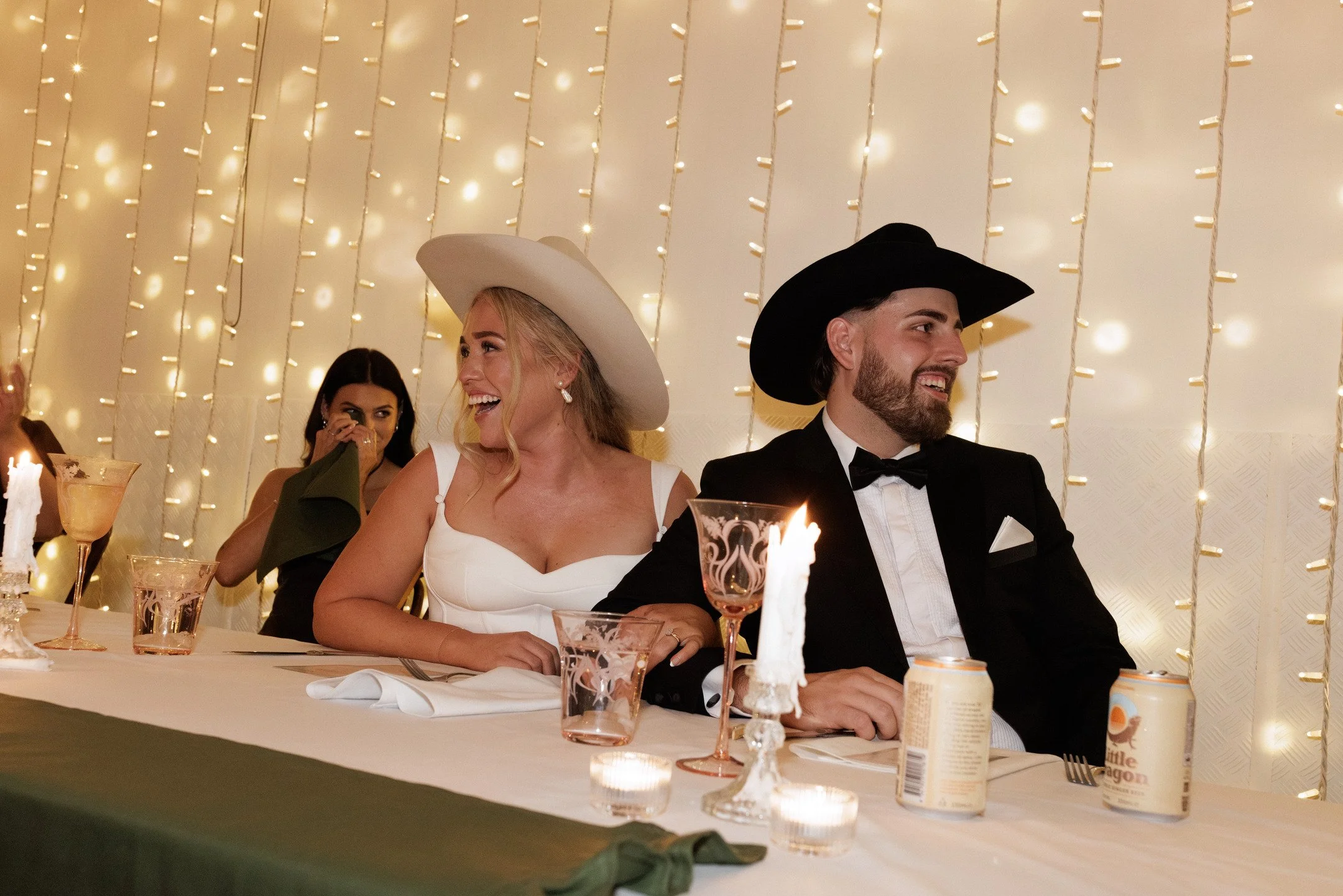 A woman in a white dress and hat and a man in a black tuxedo and hat are sitting at a decorated table, smiling and talking. There are other women in the background and string lights hanging on the wall behind them.