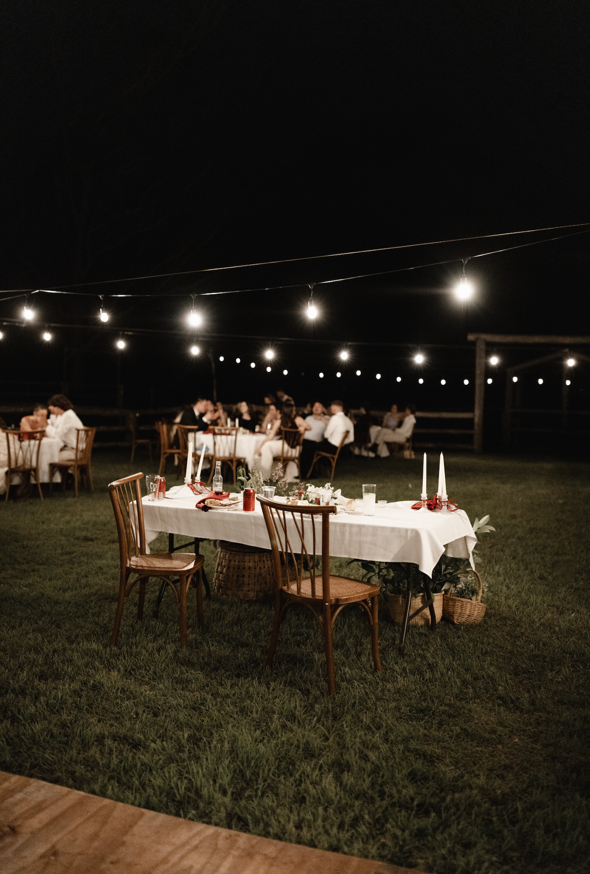 Outdoor nighttime celebration with tables set for dinner, string lights overhead, and guests seated in the background.