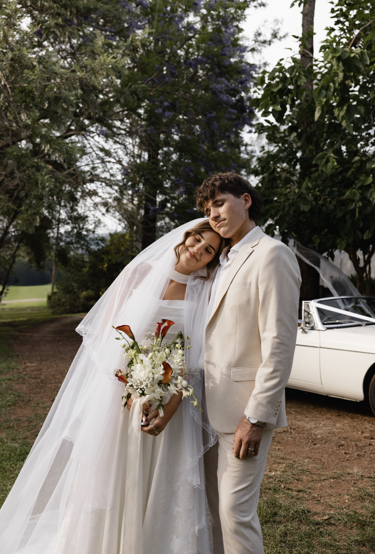 A bride and groom at their wedding outdoors, with trees and a white vintage car in the background; the bride leans her head on the groom's shoulder, holding a bouquet of flowers, both appearing happy.