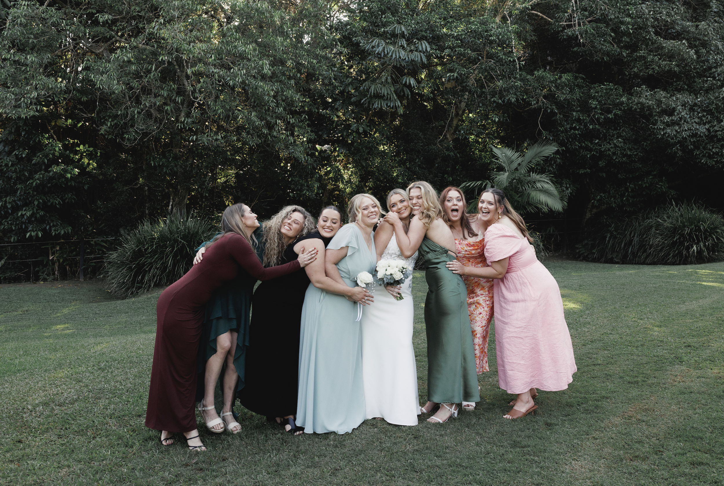 Group of women celebrating outdoors, with lush green trees in the background, some hugging and smiling, one woman in the center holding a bouquet and wearing a white dress, indicating a wedding celebration.