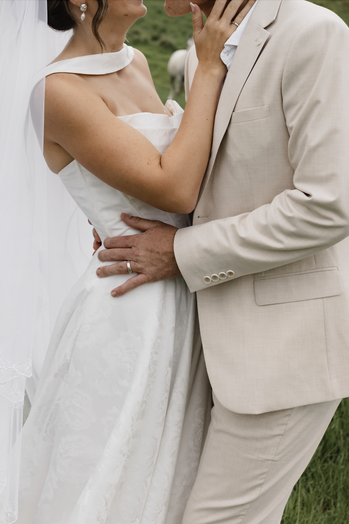 Close-up of a bride and groom embracing at their wedding, with the bride's hand touching the groom's face and the groom's hand on the bride's waist, outdoors on grass.