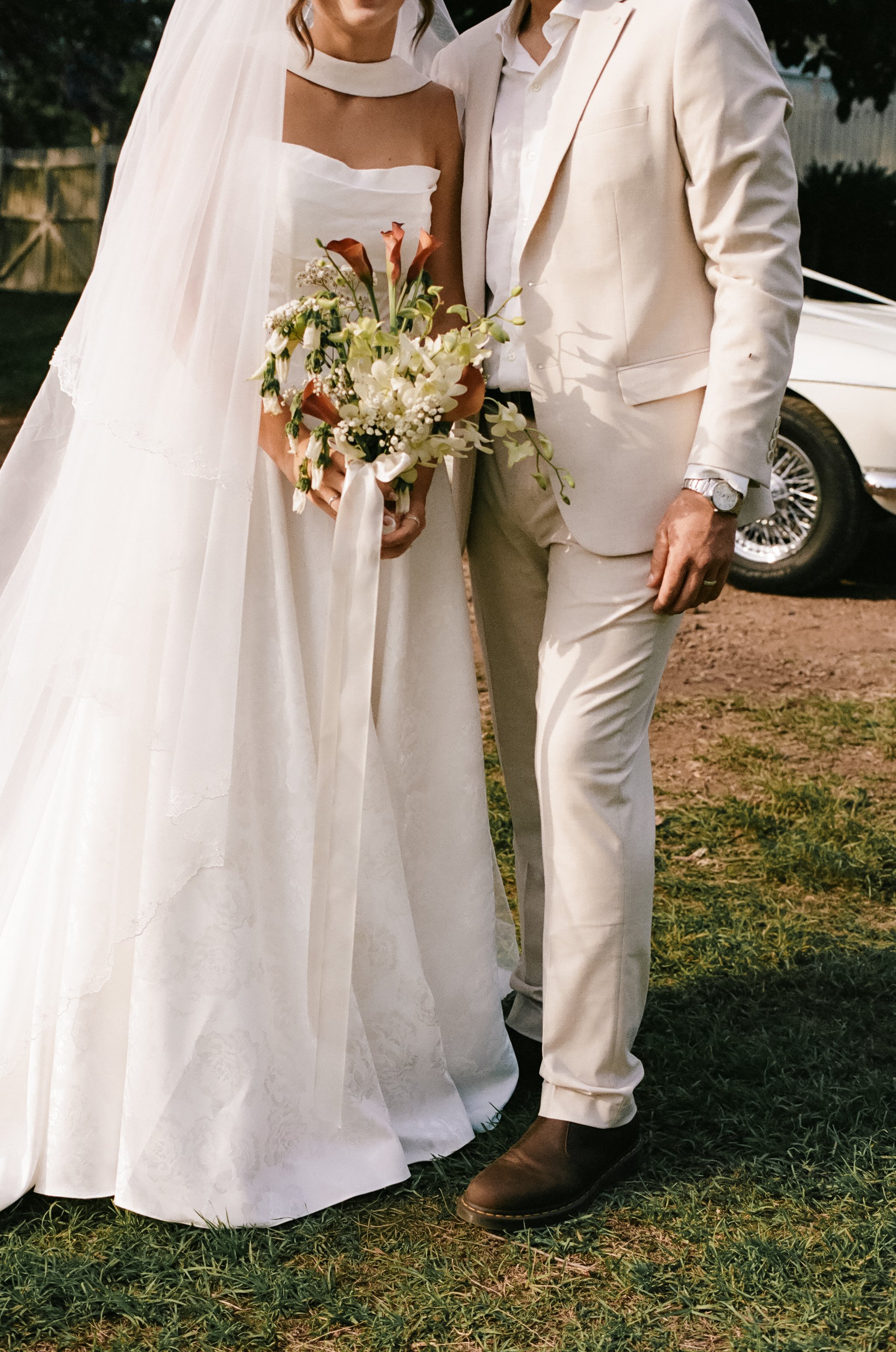 Close-up of a bride and groom standing together outdoors, with the bride holding a bouquet of flowers, and a vintage white car in the background.