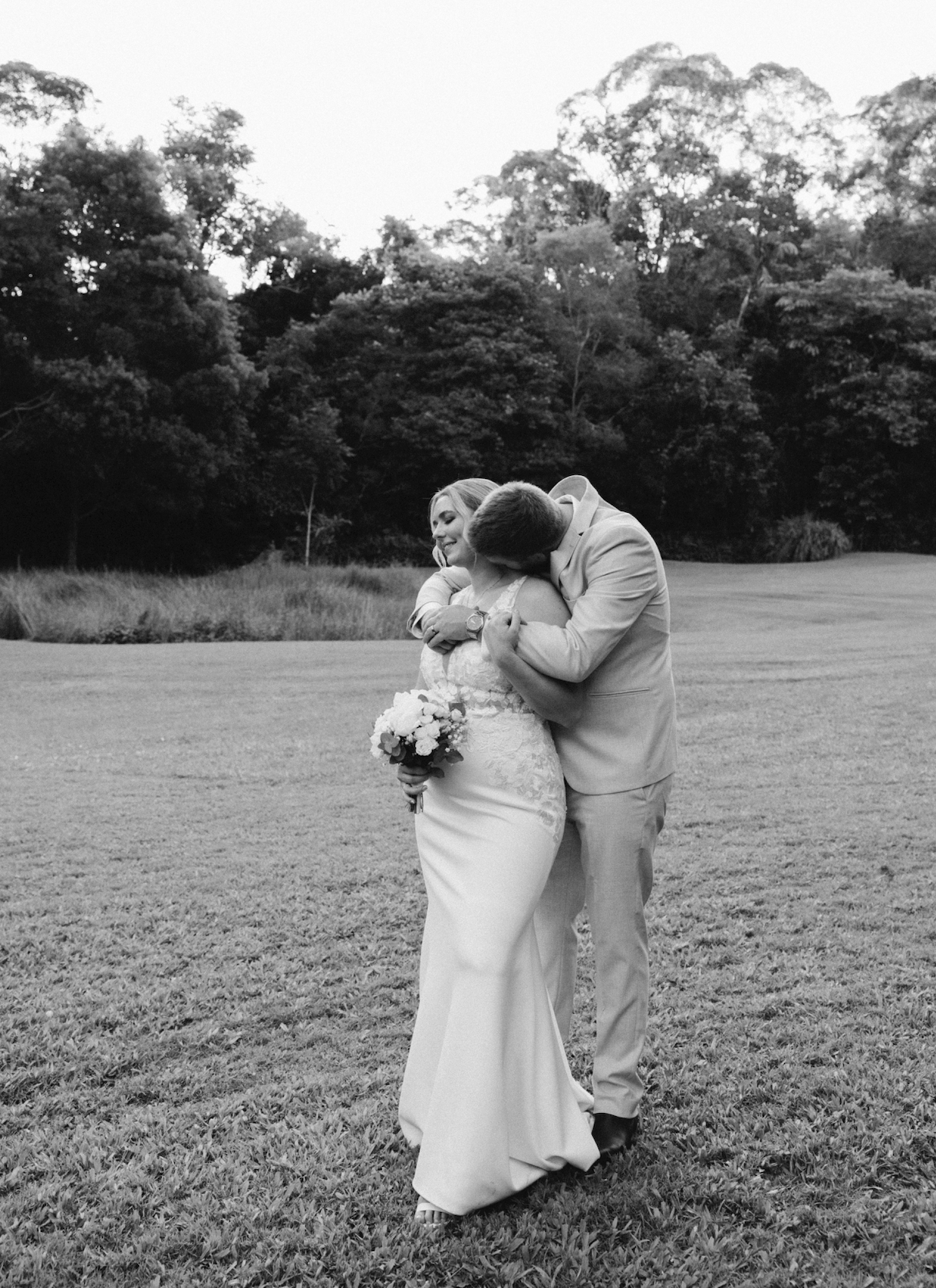 Black-and-white photo of a couple on their wedding day, embracing outdoors in a grassy field with trees in the background. The bride is holding a bouquet and smiling, while the groom is kissing her on the cheek.