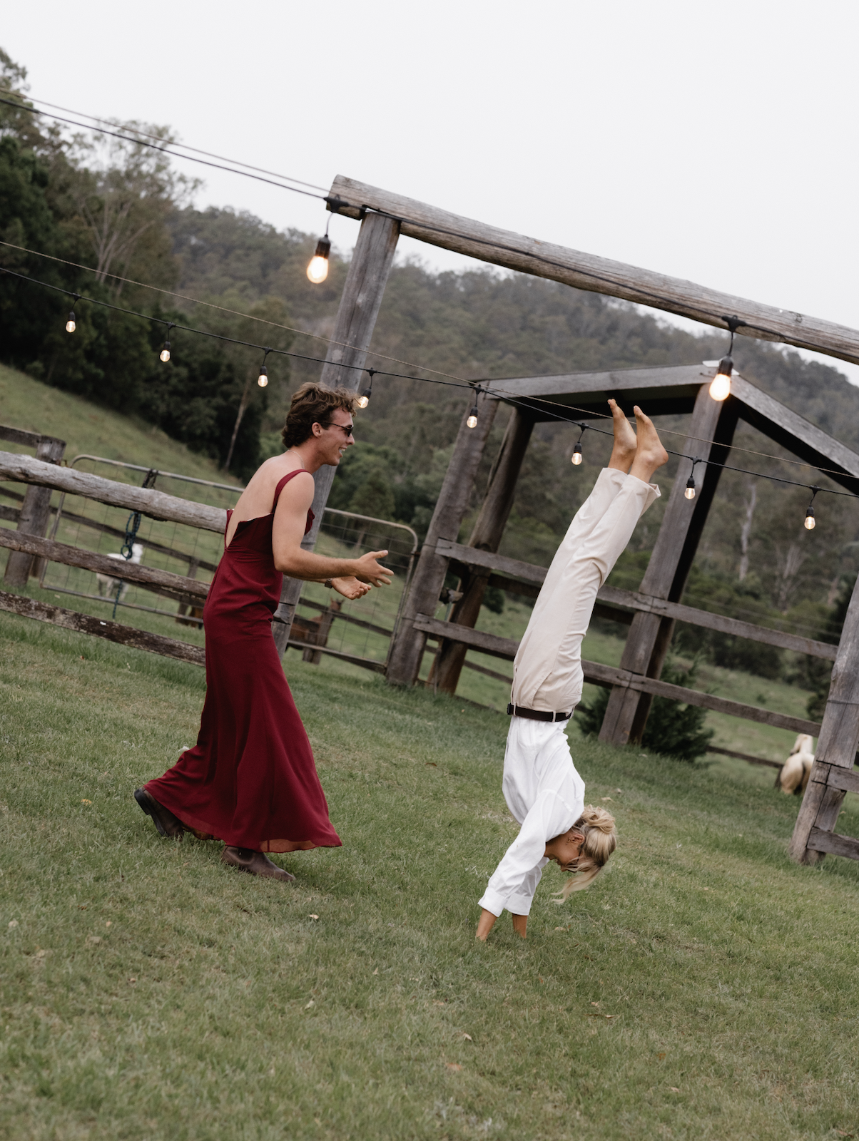 A woman in a red dress is laughing and watching a girl doing a handstand in a grassy outdoor area with string lights and wooden structures.