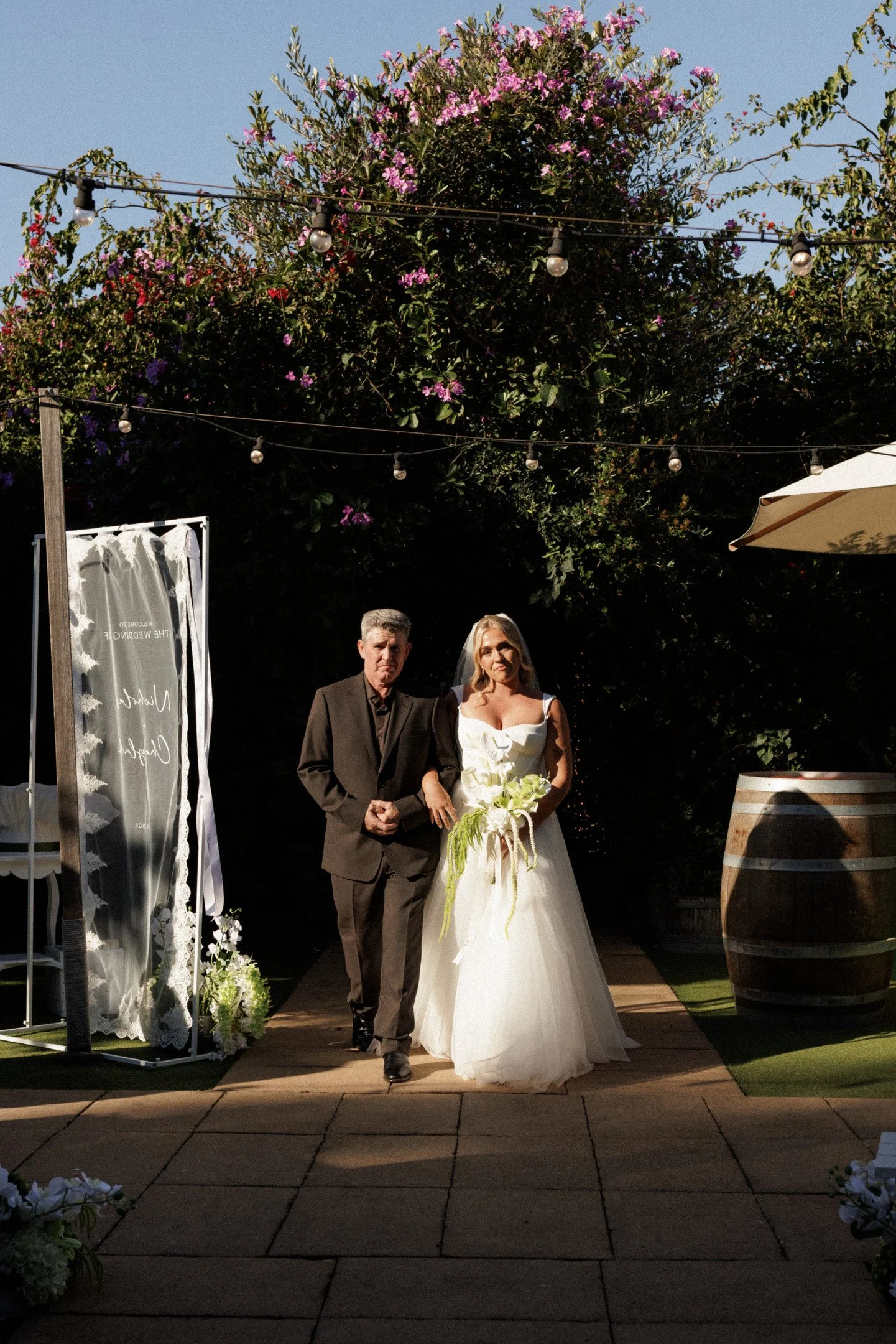 A bride and an older man, likely her father, walking down the aisle at an outdoor wedding ceremony under string lights, with a large flowering tree in the background.