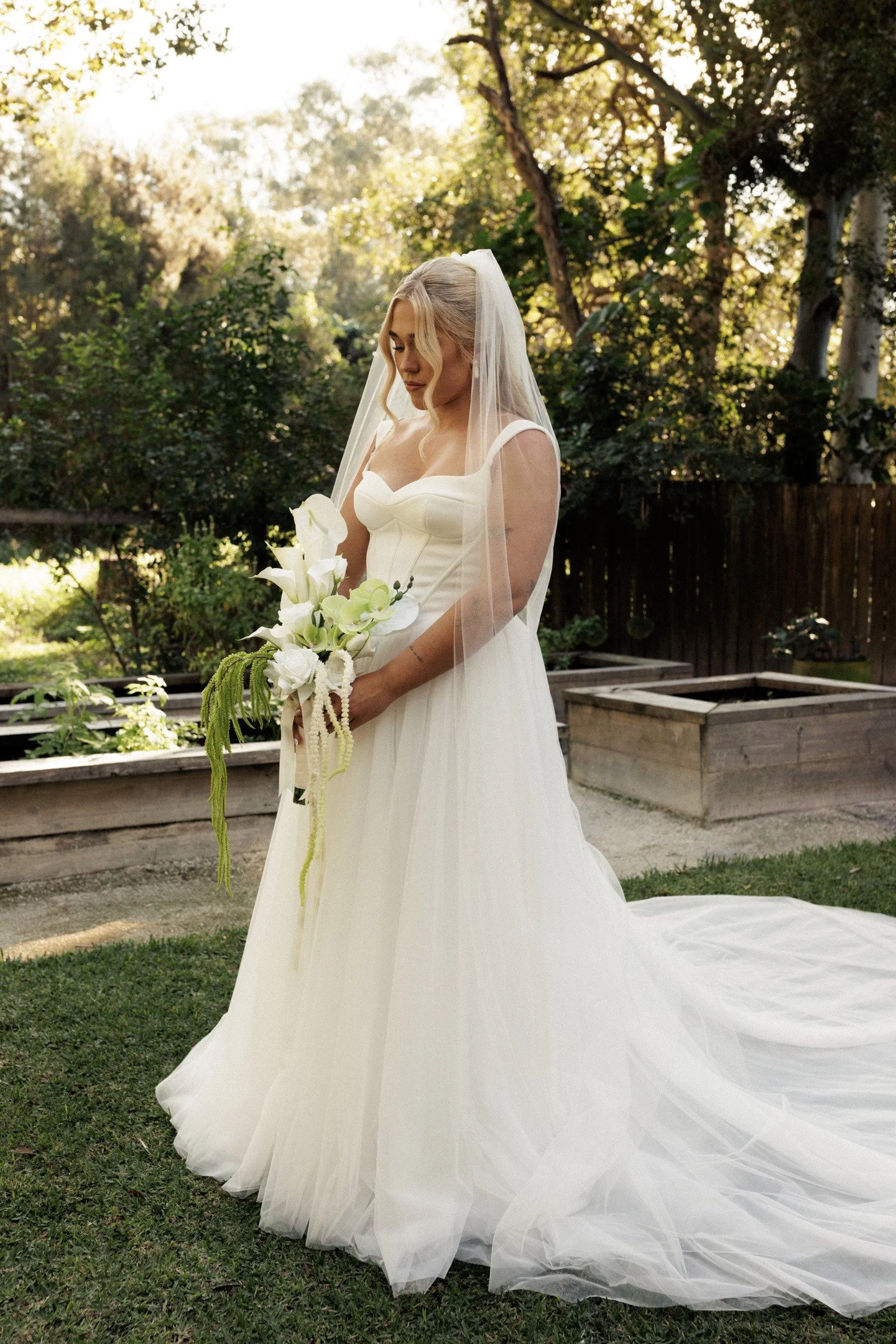 A bride in a white wedding dress with a long train and sheer veil, holding a bouquet of white flowers, standing outdoors in a garden with trees and wooden planters.