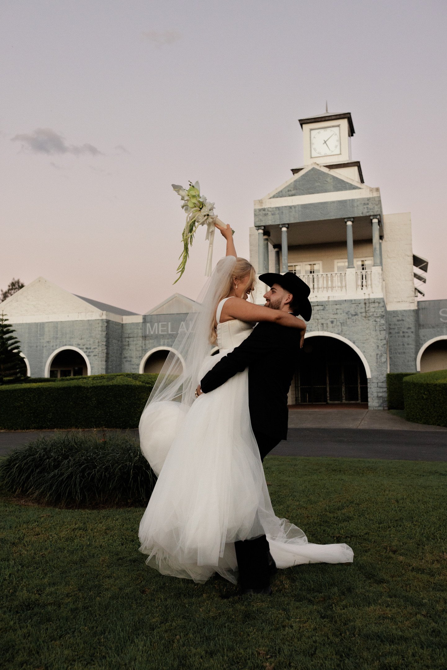 Bride in a white wedding gown being lifted by groom in a black suit and cowboy hat in front of a building with columns and a clock tower, holding a bouquet in the air, during sunset.