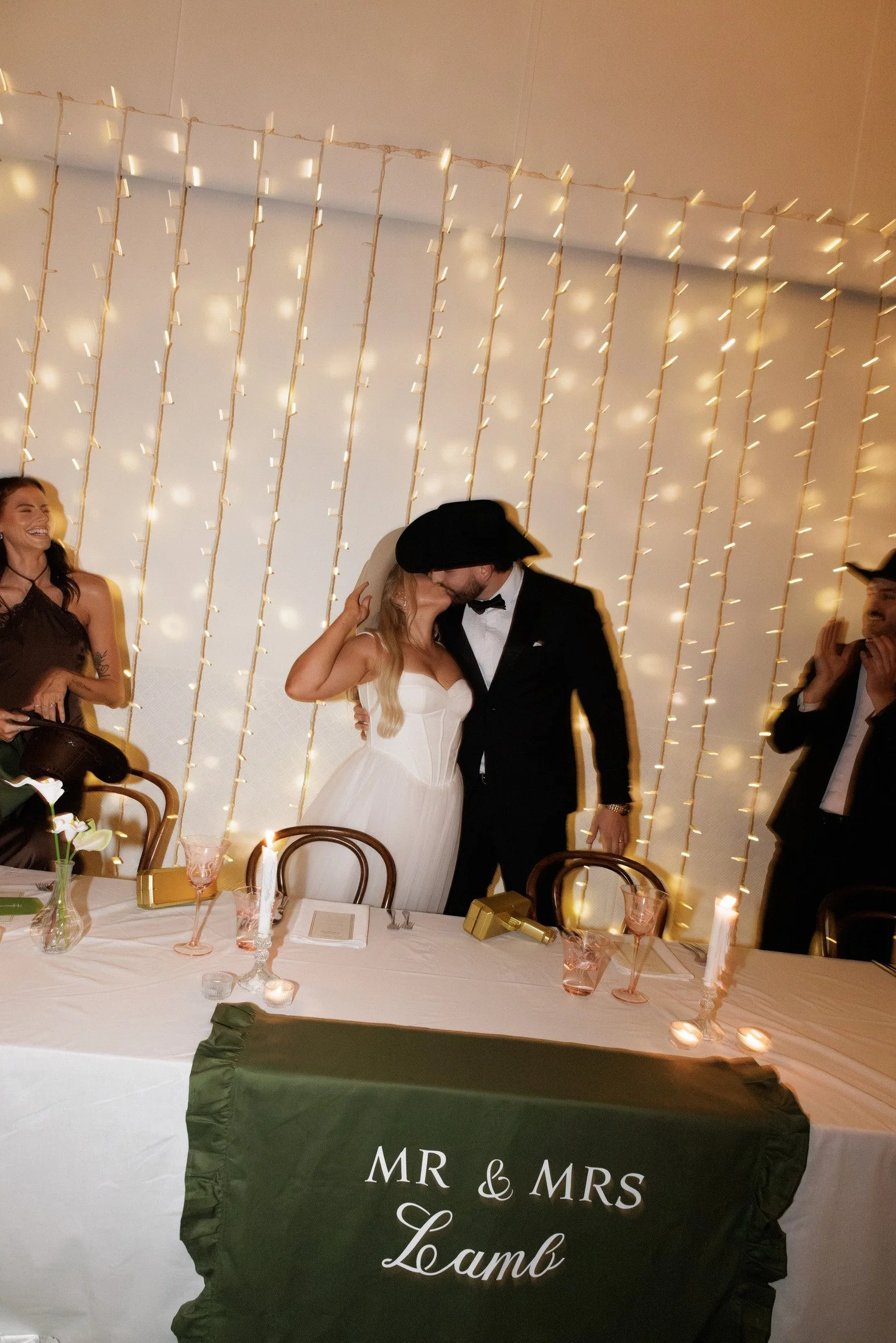 A wedding reception with a bride and groom kissing in front of a decorated backdrop of string lights. Guests are seated at a table with candles, flowers, and glasses. A black banner on the table reads 'MR & MRS Lamb'.