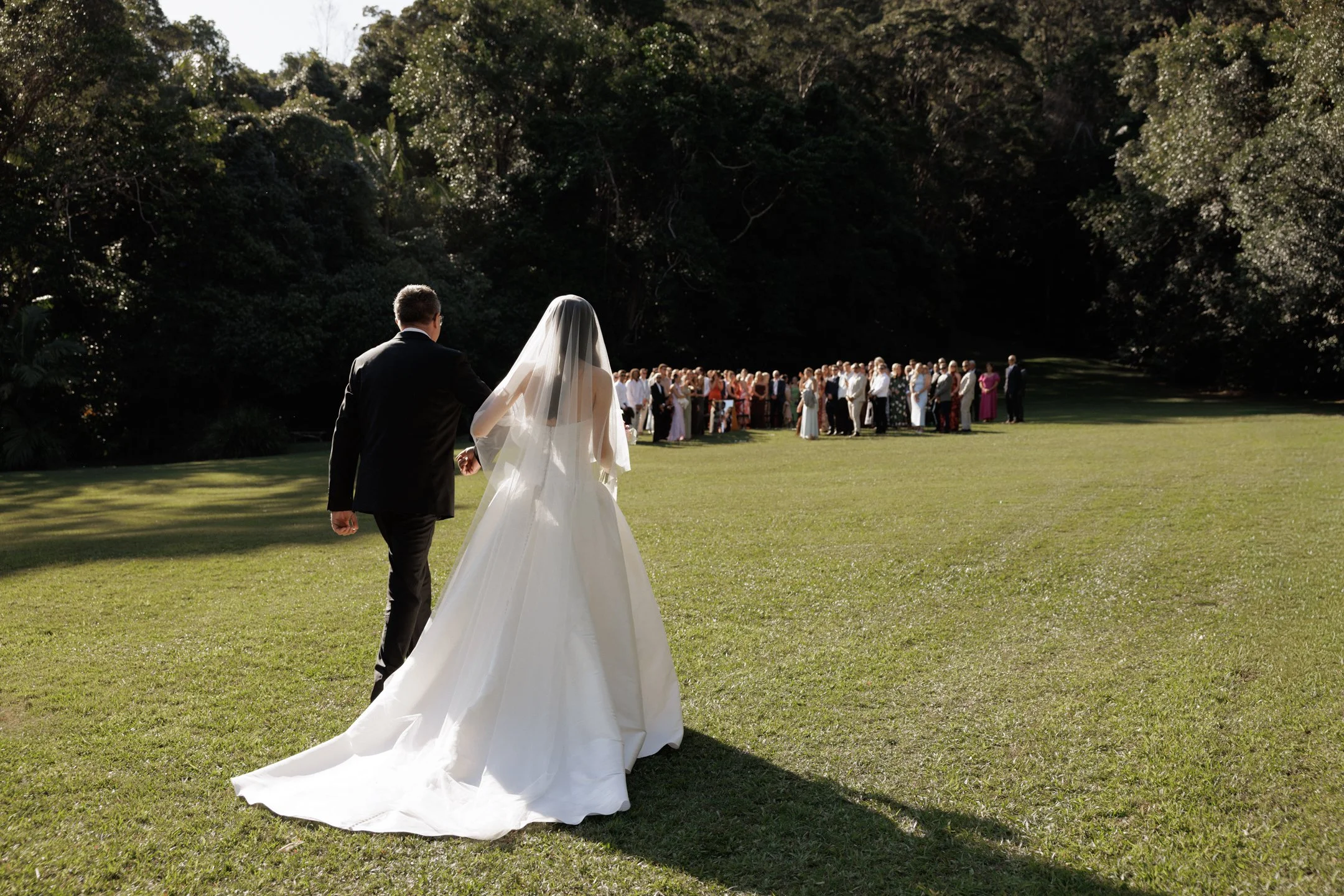 A bride and groom walk together on a grassy field during an outdoor wedding ceremony, with guests seated in the background.