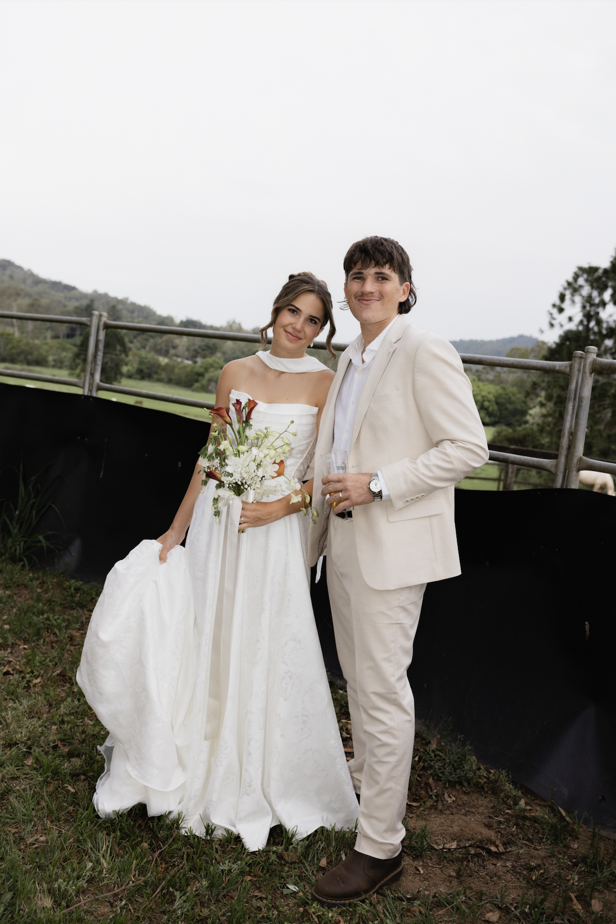 A bride and groom stand outdoors on a cloudy day, posing for a photo. The bride is in a white wedding gown holding a bouquet of white and reddish flowers, and the groom is in a light beige suit holding a drink. They are smiling and standing in front 