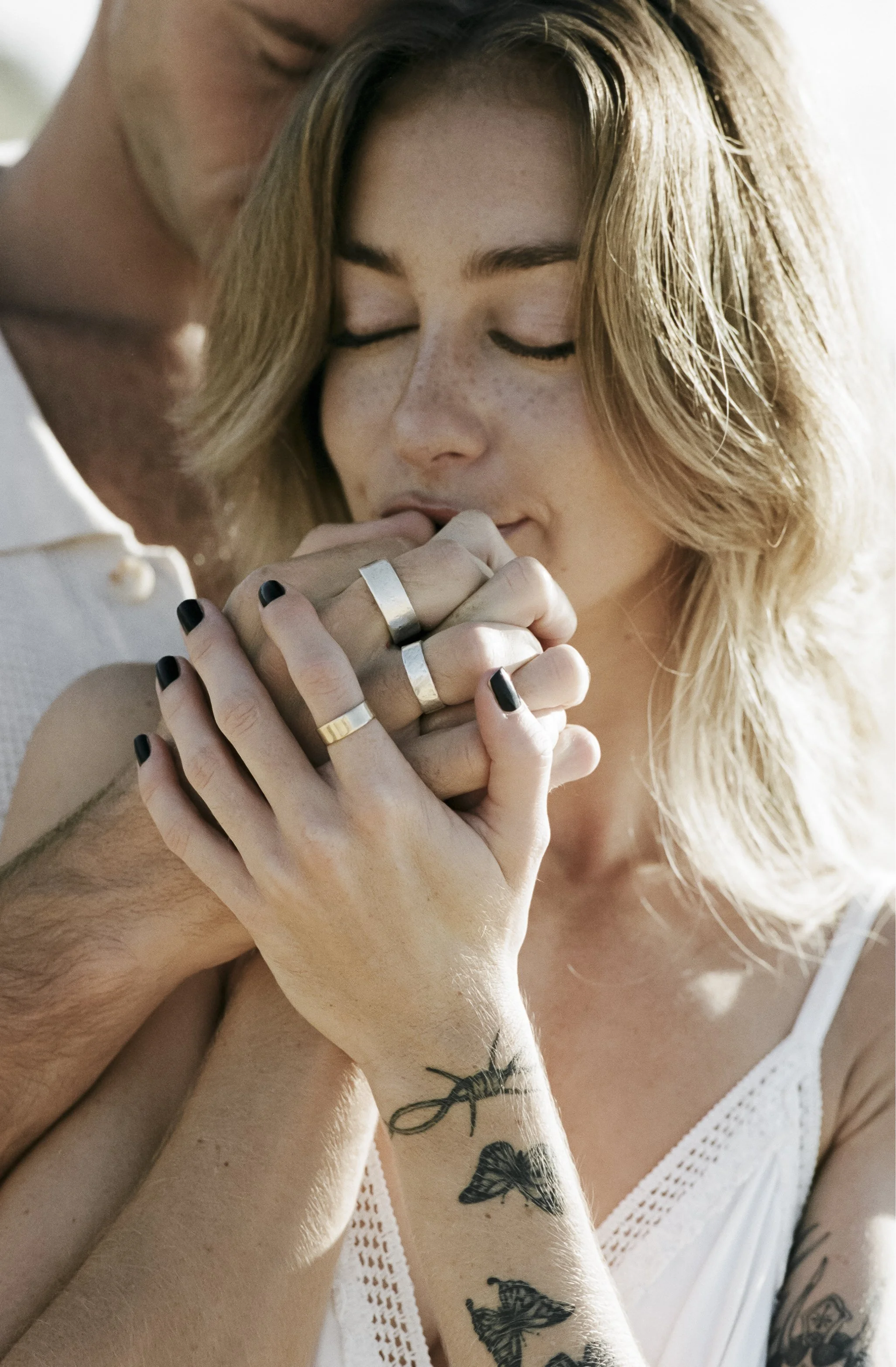 A couple holding hands with rings, close-up of woman with tattoos and closed eyes, embracing affectionately.