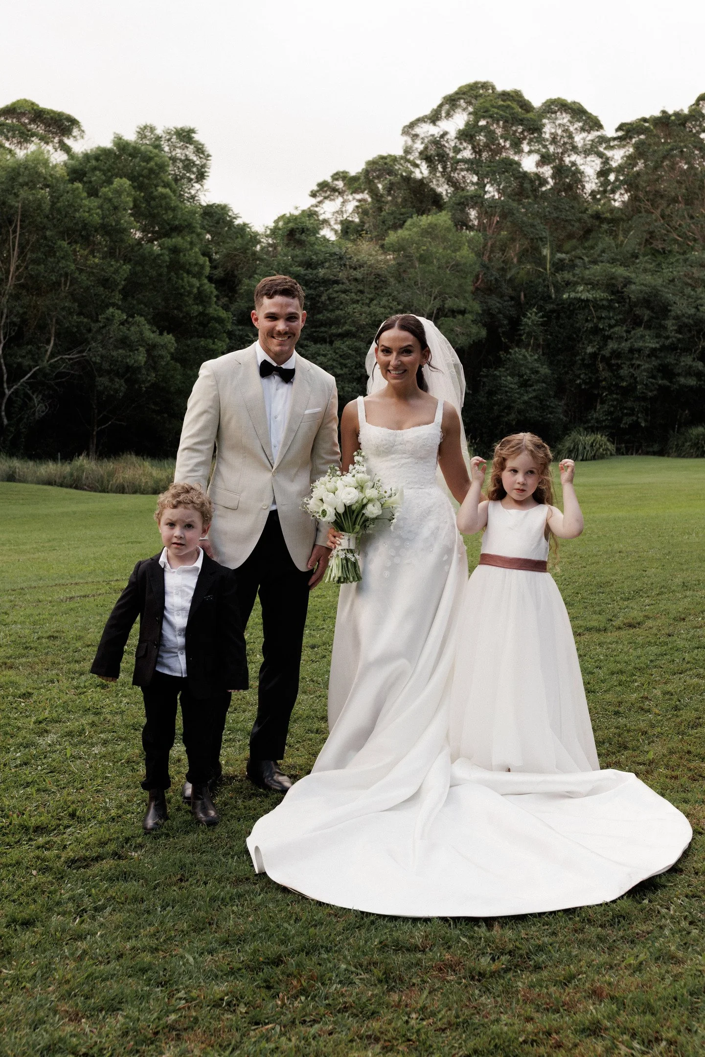 A bride and groom standing outdoors on a grassy area with two young children, a boy and a girl, near them. The groom is wearing a beige tuxedo with a black bow tie, and the bride is in a white wedding dress holding a bouquet of white flowers. The boy