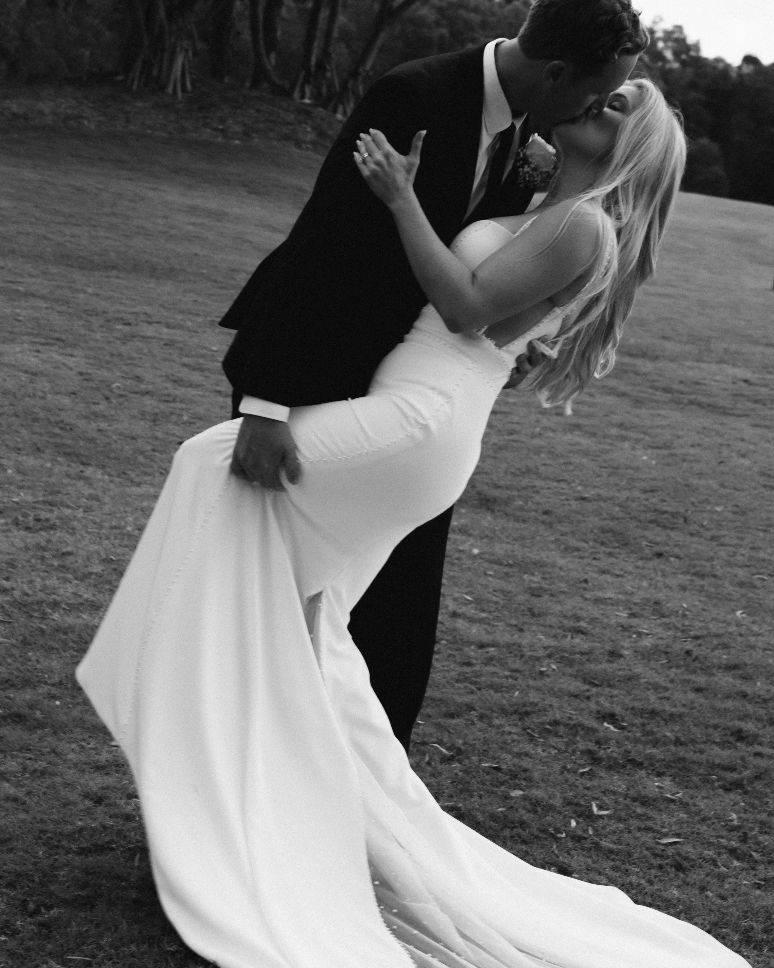 A black-and-white photo of a couple in wedding attire sharing a kiss outdoors, with the man holding the woman as she leans back.