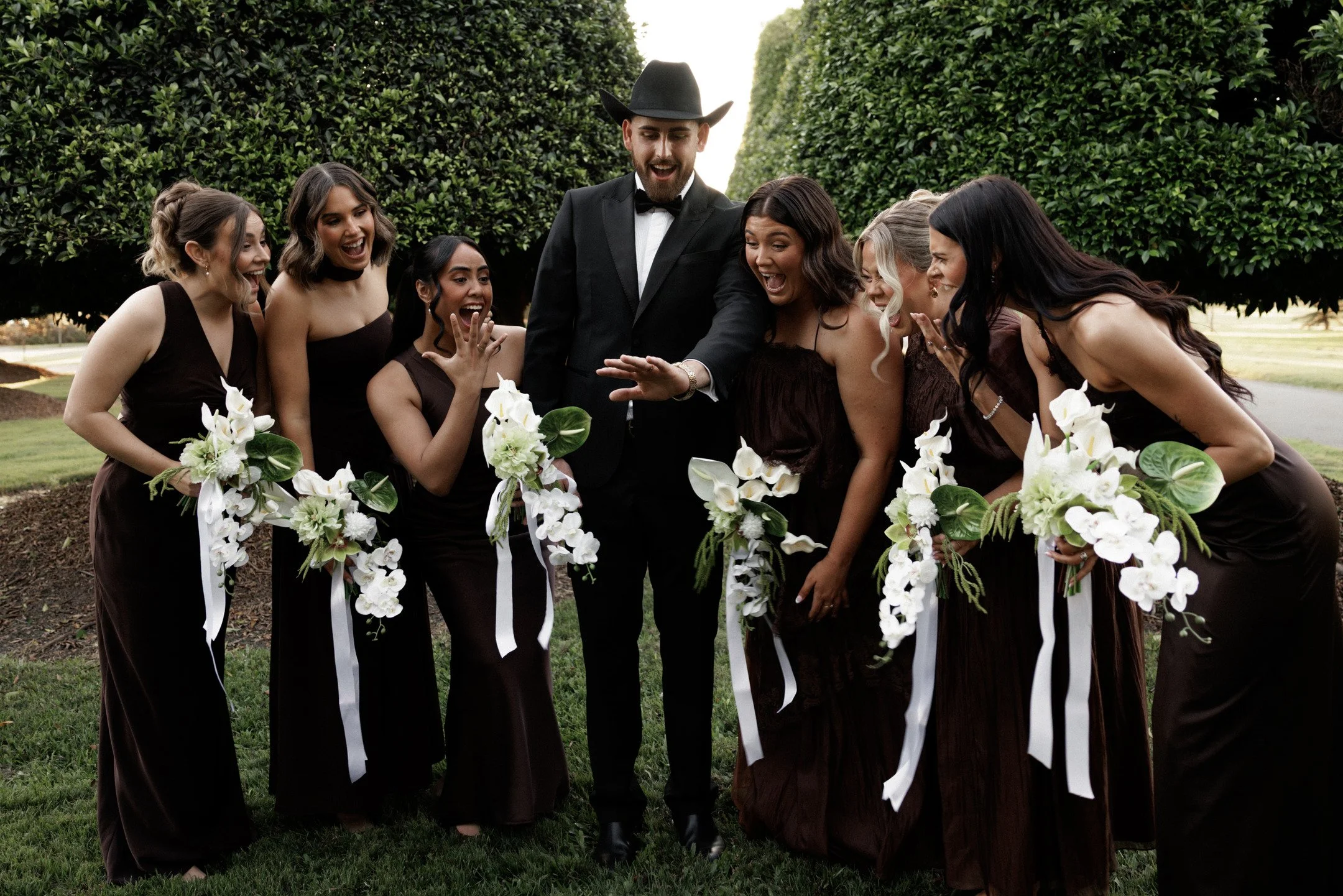 A group of women dressed in matching dark brown dresses with white ribbons and holding white floral bouquets surrounding a man in a black suit and cowboy hat, outdoors with greenery in the background.