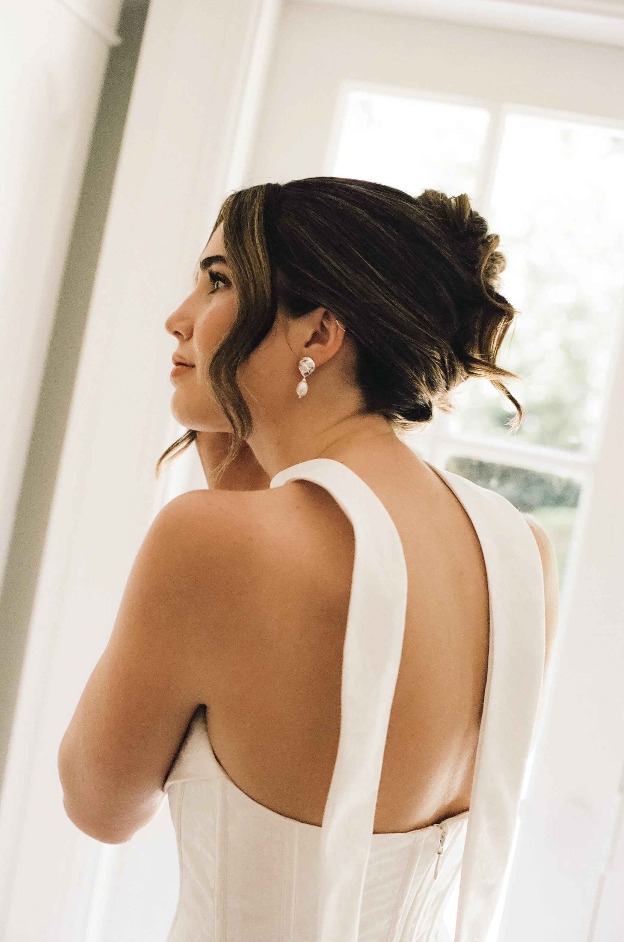 A woman with dark hair styled in an updo, wearing a white dress with off-shoulder straps and pearl earrings, standing indoors near a window.