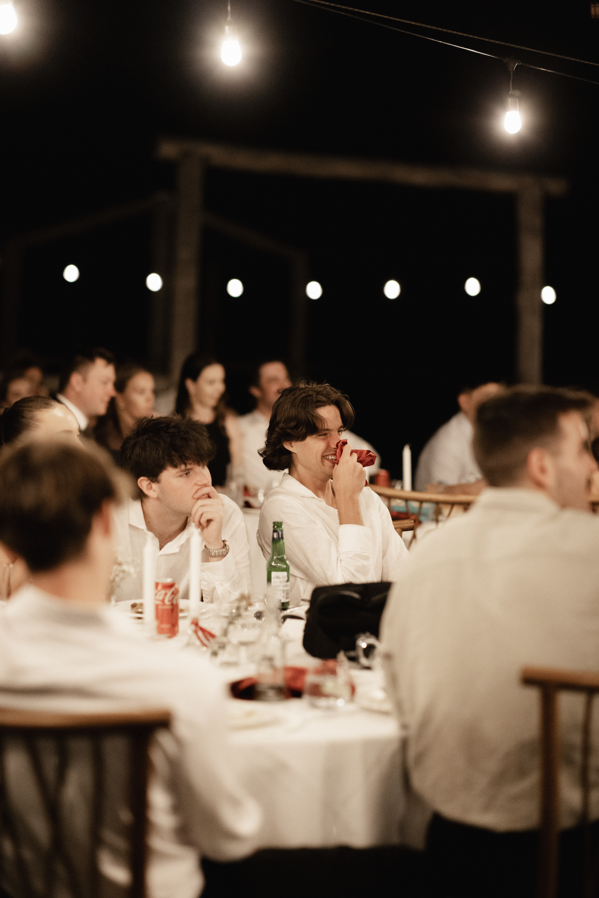 People seated at a dinner table at night, with string lights overhead, some smiling and laughing.