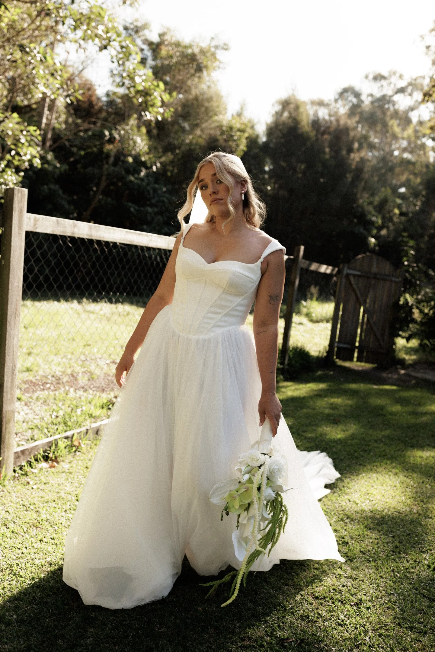 A bride in a white wedding gown holding a bouquet of white flowers outdoors in a garden with trees and a wooden fence, taken during the daytime.