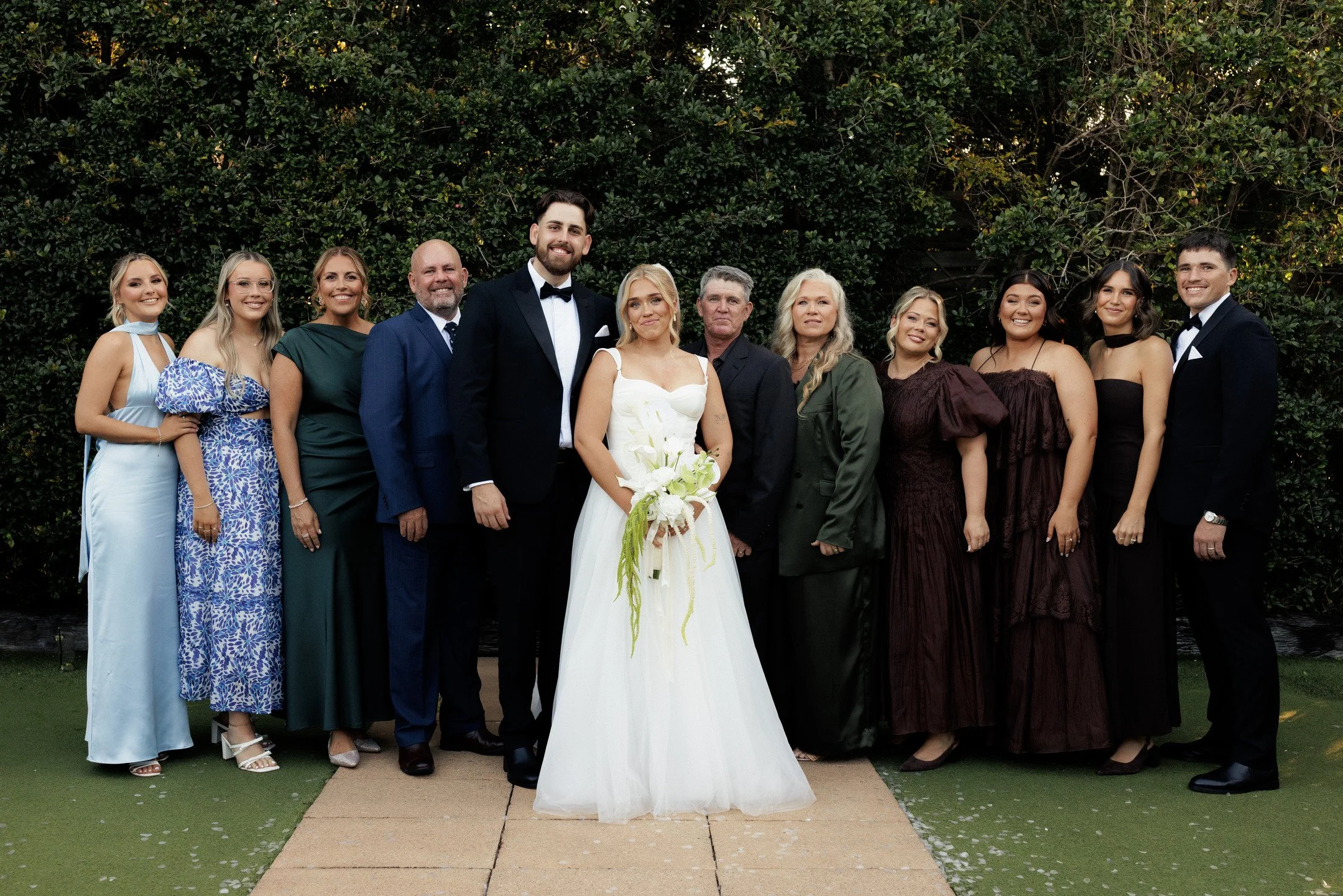 A group of people dressed in formal attire, including a bride in a white wedding gown holding a bouquet, and others in suits and dresses, standing outdoors in front of a hedge, celebrating a wedding.