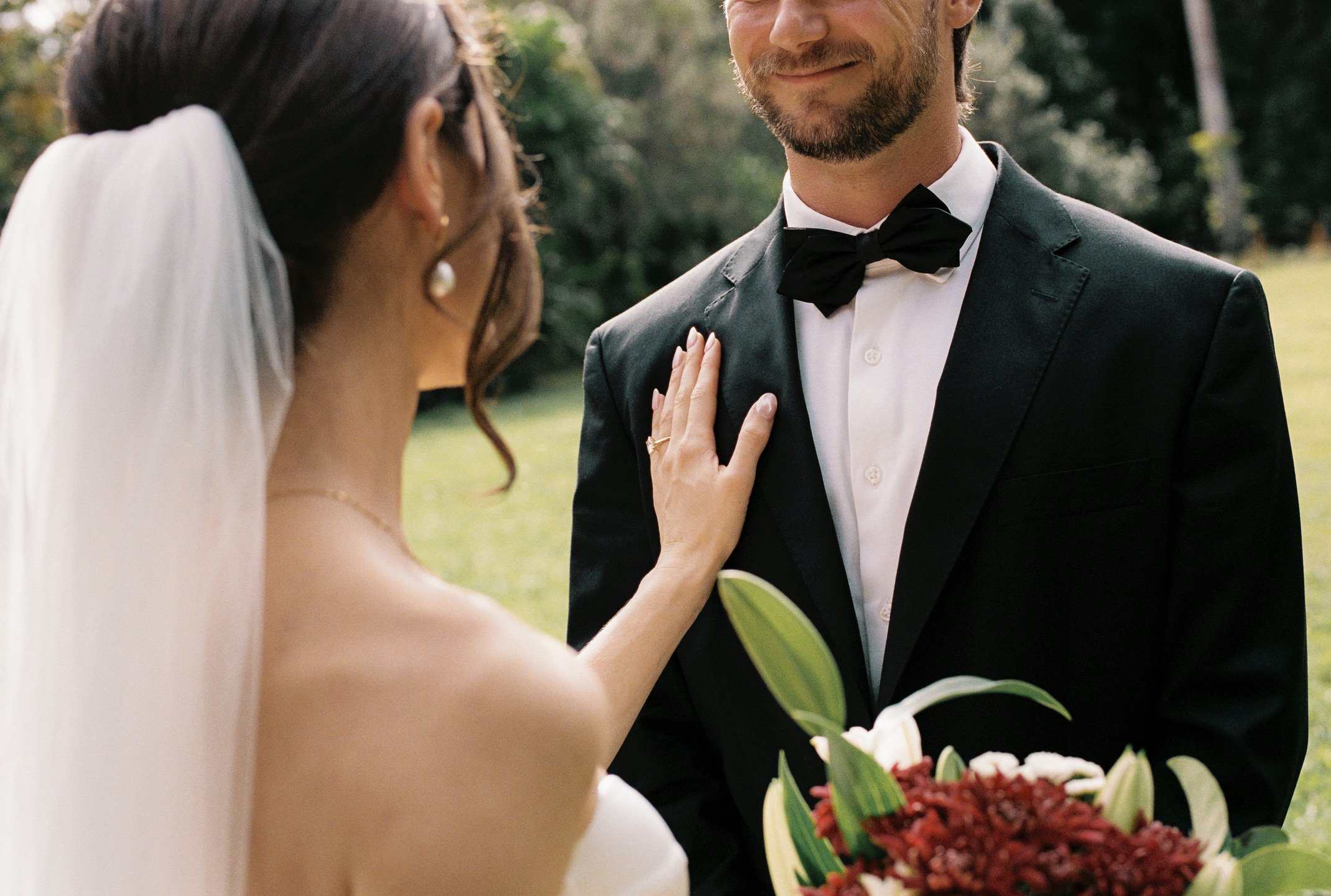 A bride and groom during a wedding ceremony outdoors, with the bride reaching out to touch the groom's chest. The bride is wearing a white dress with a veil, and the groom is in a black tuxedo with a bow tie, holding a bouquet of flowers.