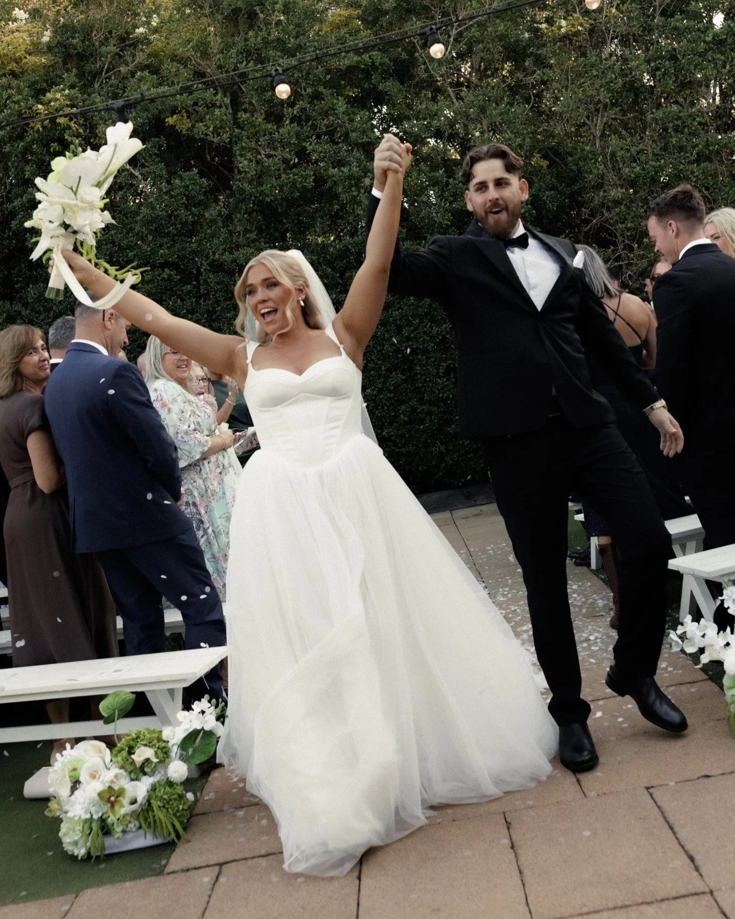 A bride and groom dancing at their wedding. The bride is wearing a white wedding dress and holding a bouquet, while the groom is in a black tuxedo. Guests are watching and smiling in the background.