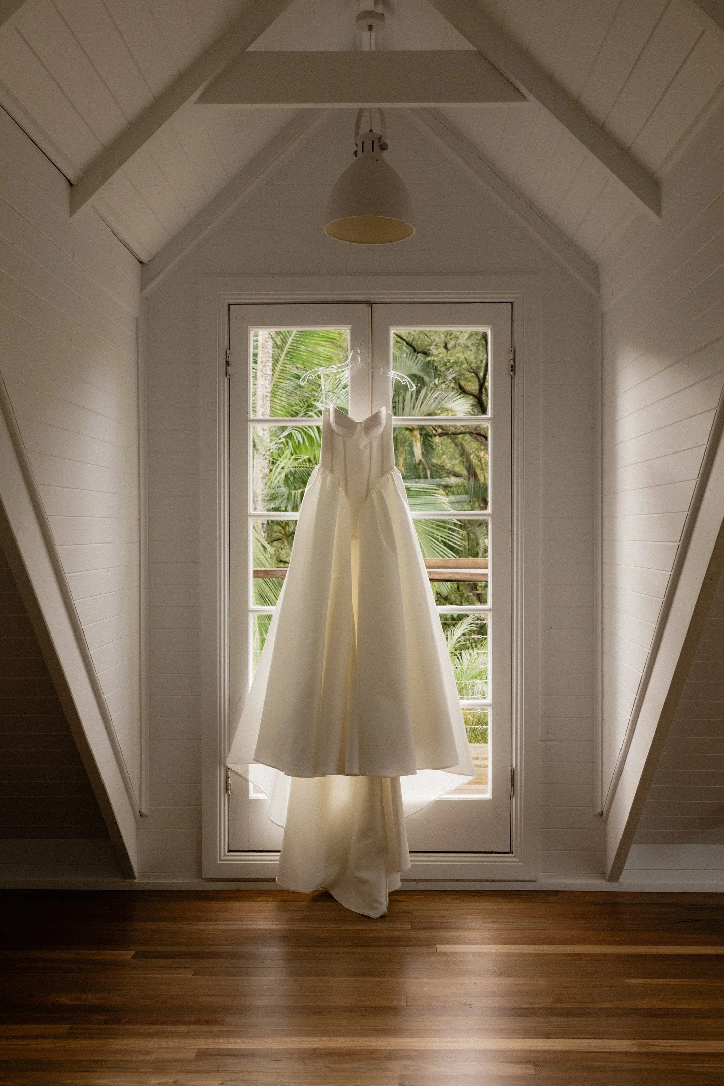 Wedding dress hanging in front of a glass door with a view of green trees outside, inside a room with wooden floor.