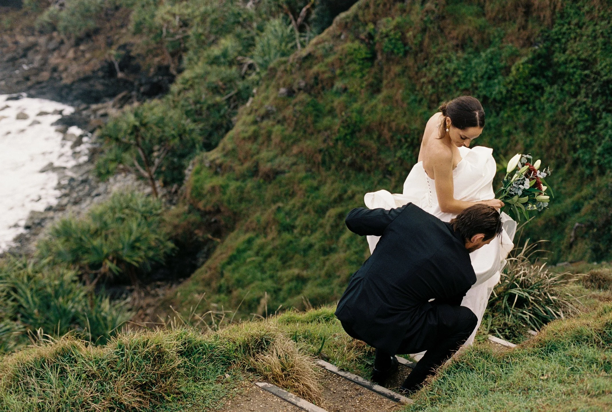 A man proposing marriage to a woman on a grassy hillside near a river, with the woman holding a bouquet of flowers and looking down at him.