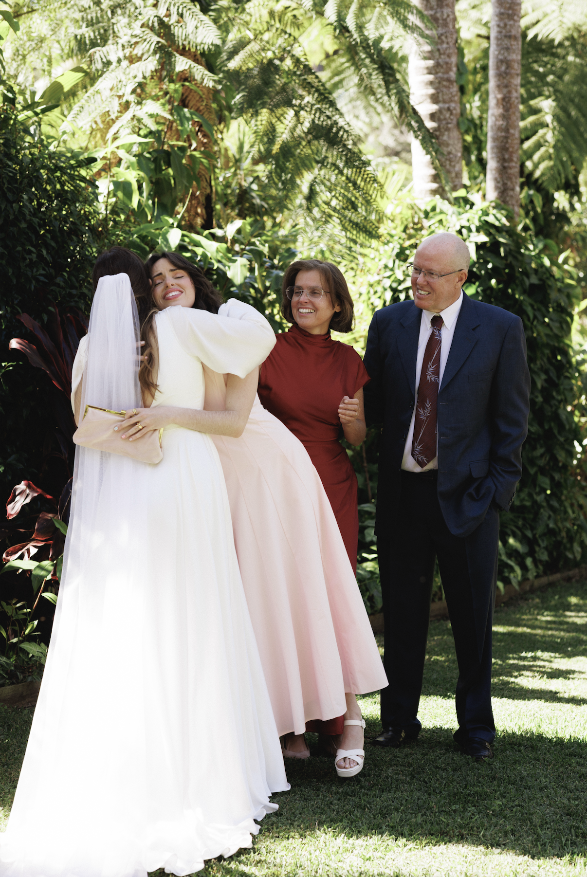 People hugging and smiling outdoors in a lush garden during daytime, including a bride in a white gown and veil, a woman in a pink dress, a woman in red, and a man in a suit.