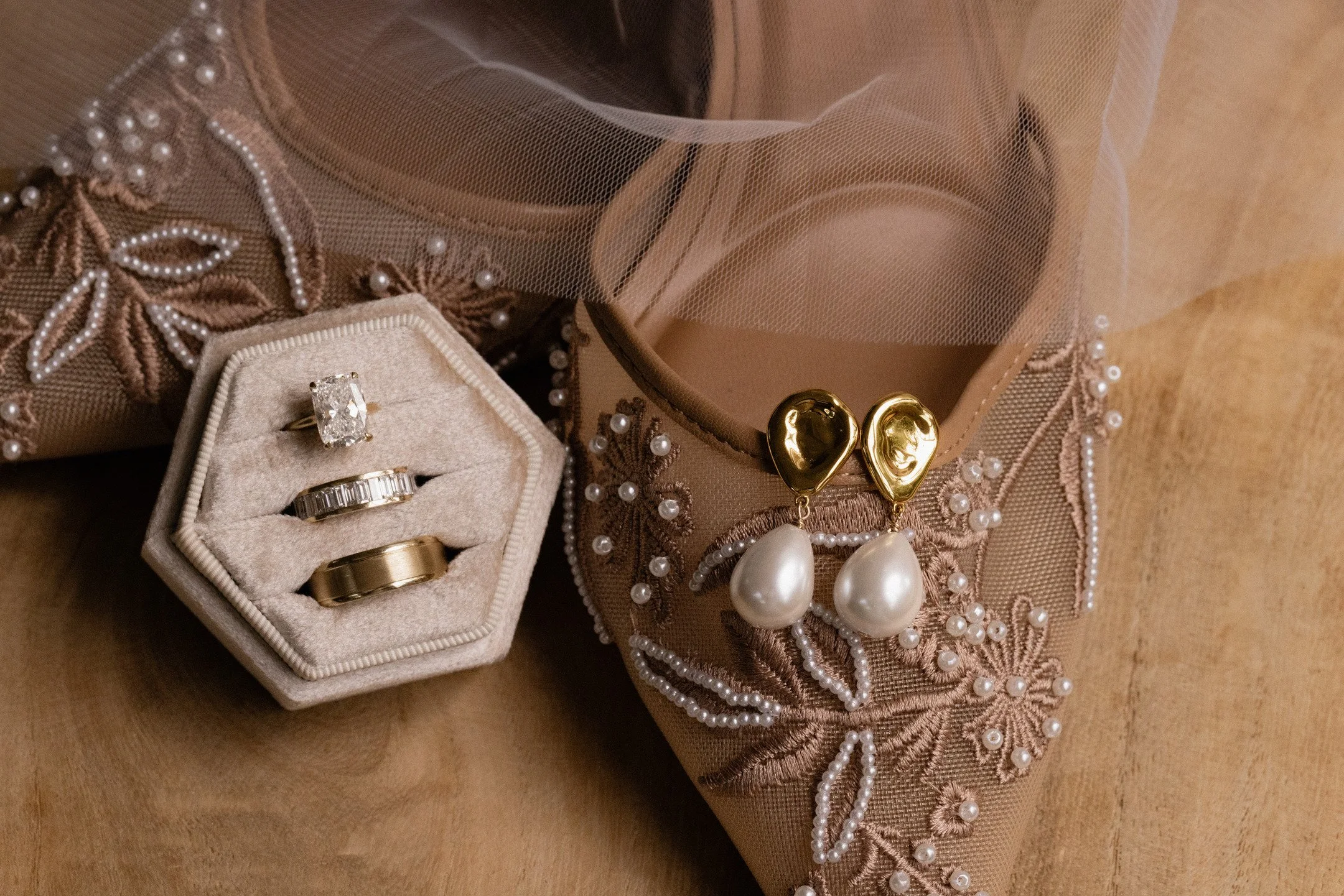 Jewelry box containing engagement ring, wedding bands, earrings with large pearls, and a necklace with pearls, all set on a decorative fabric surface with beads and embroidery.