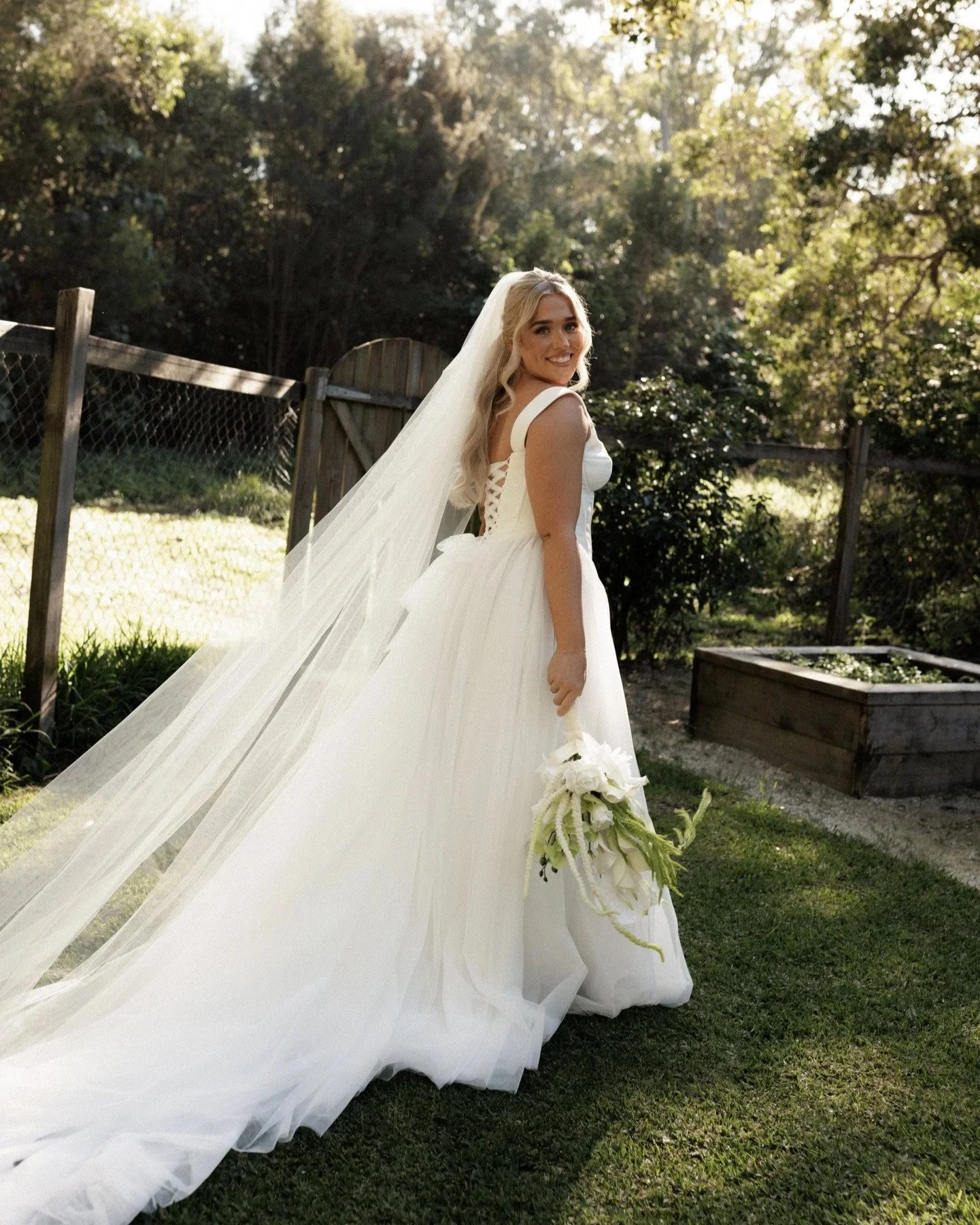 A smiling woman in a white wedding dress standing outdoors, holding a bouquet of white flowers, with a wooden fence and trees in the background.