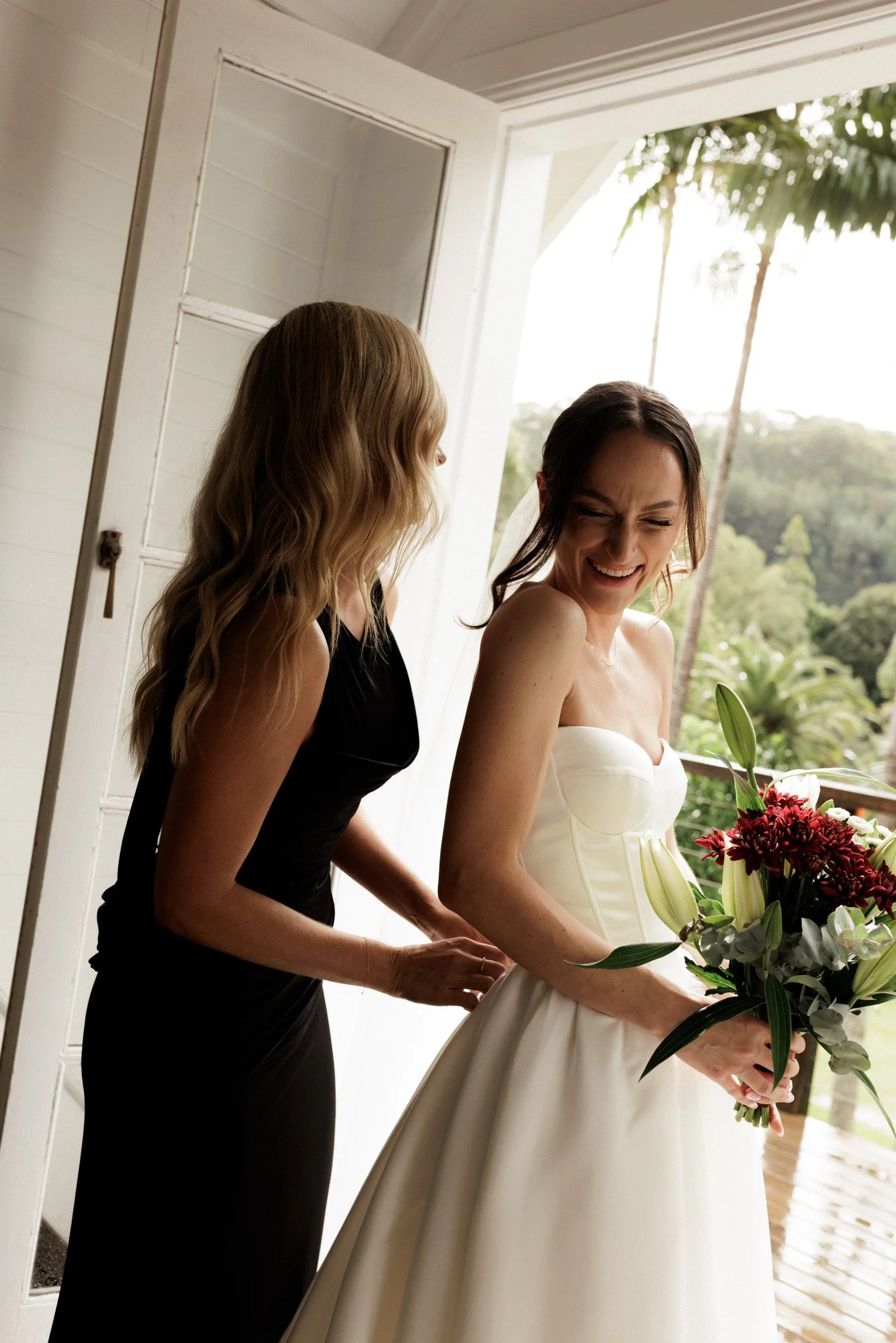 Two women, one in a black dress and the other in a white wedding gown, standing near an open door on a porch, with greenery and palm trees in the background, holding a bouquet of flowers.