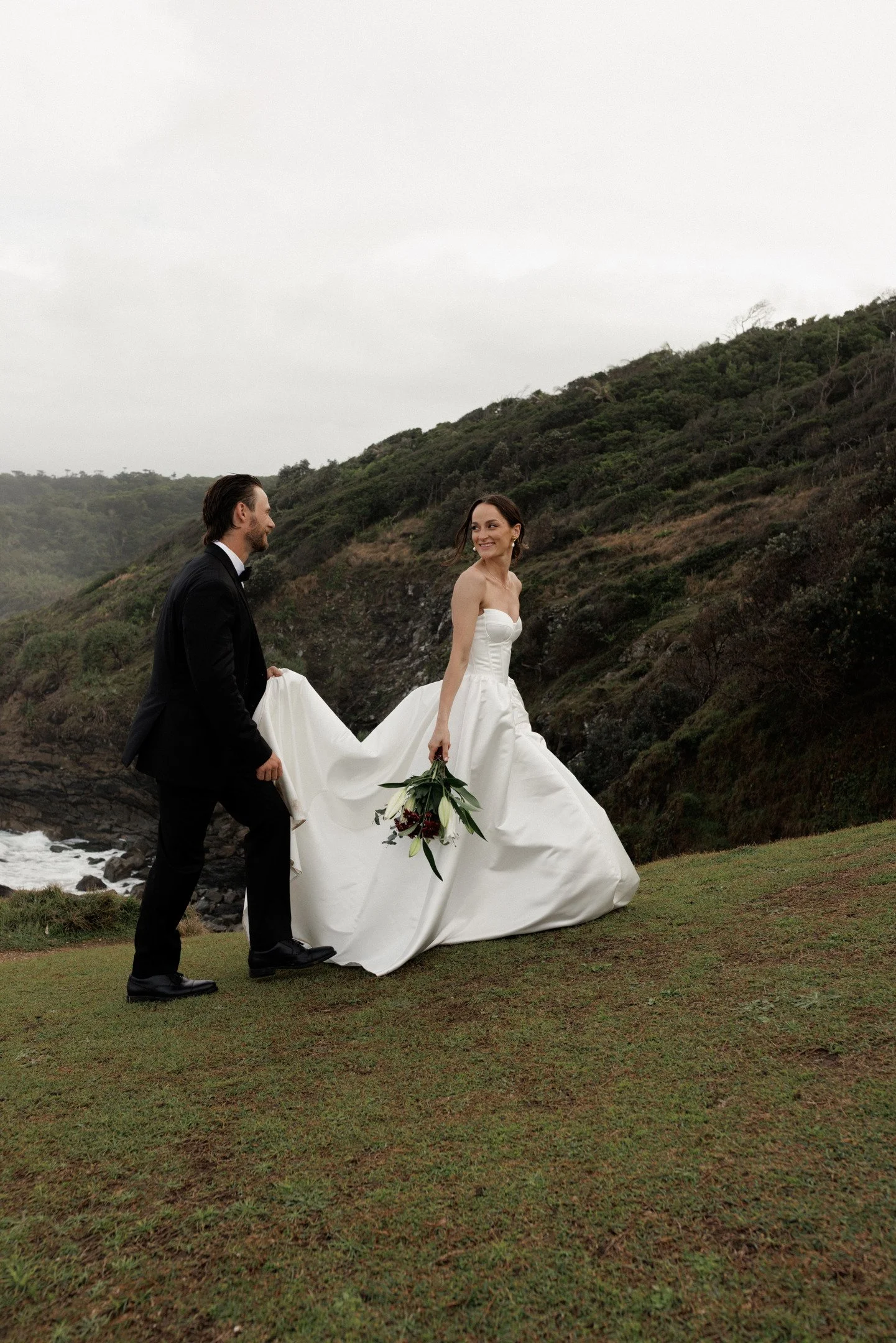A bride in a white wedding gown walking on grass by the coast, smiling and holding a bouquet, with a groom in a black tuxedo walking beside her, against a backdrop of green hillside and cloudy sky.
