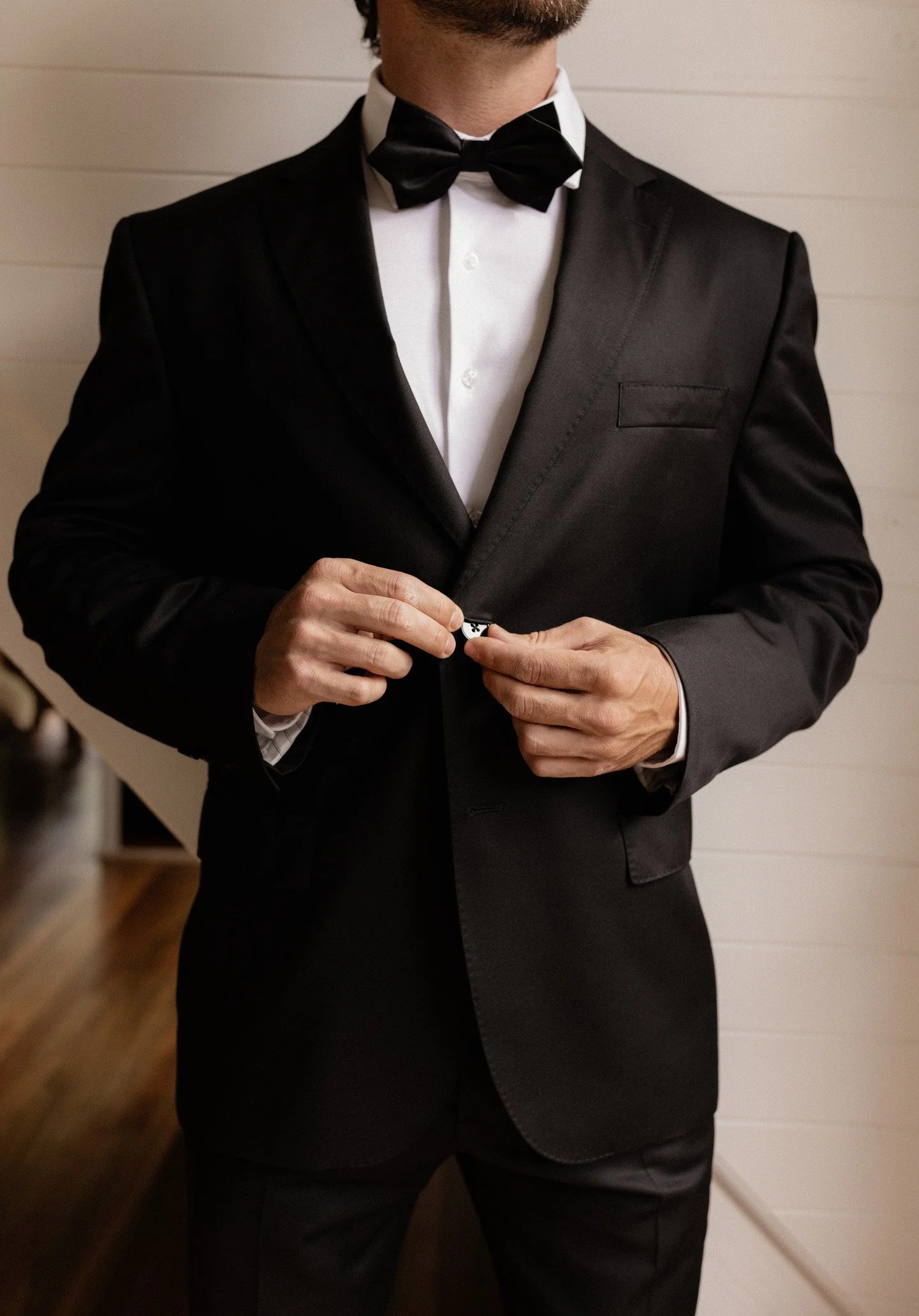 A man wearing a black tuxedo with a white dress shirt and a black bow tie, adjusting his cufflink.