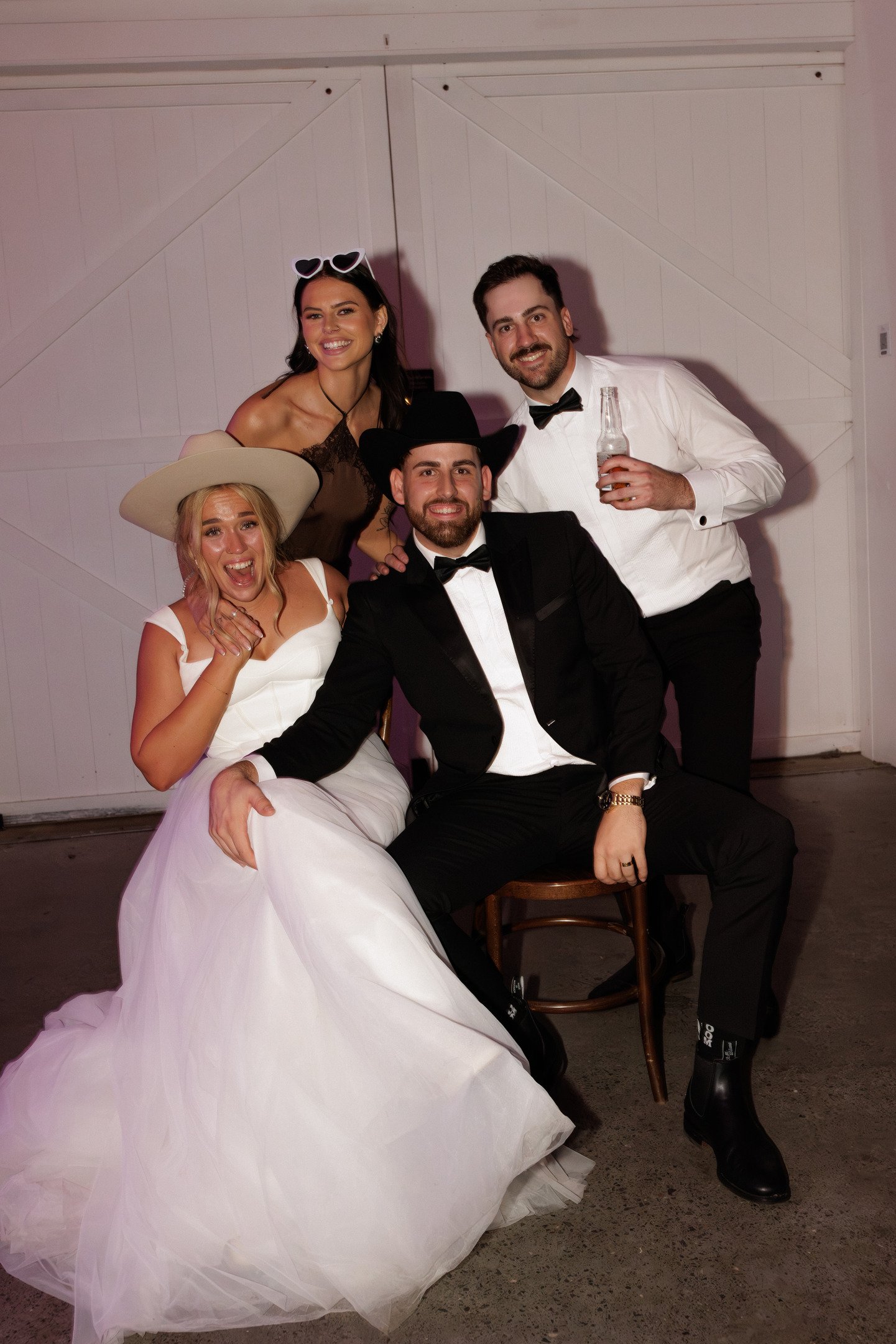 Group of four people at a celebration, two men in tuxedos, one woman in a white dress and large hat, and one woman in a brown dress with heart-shaped sunglasses, smiling and posing in front of white panel walls.
