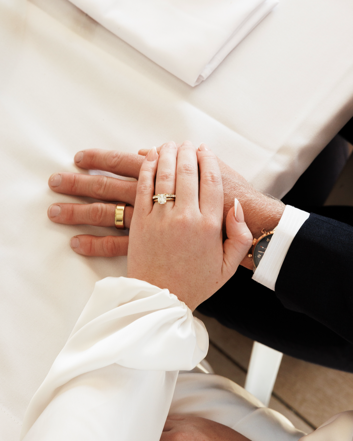 Close-up of a newlywed couple's hands, showing wedding rings, with the bride's hand resting on the groom's hand on a white table.