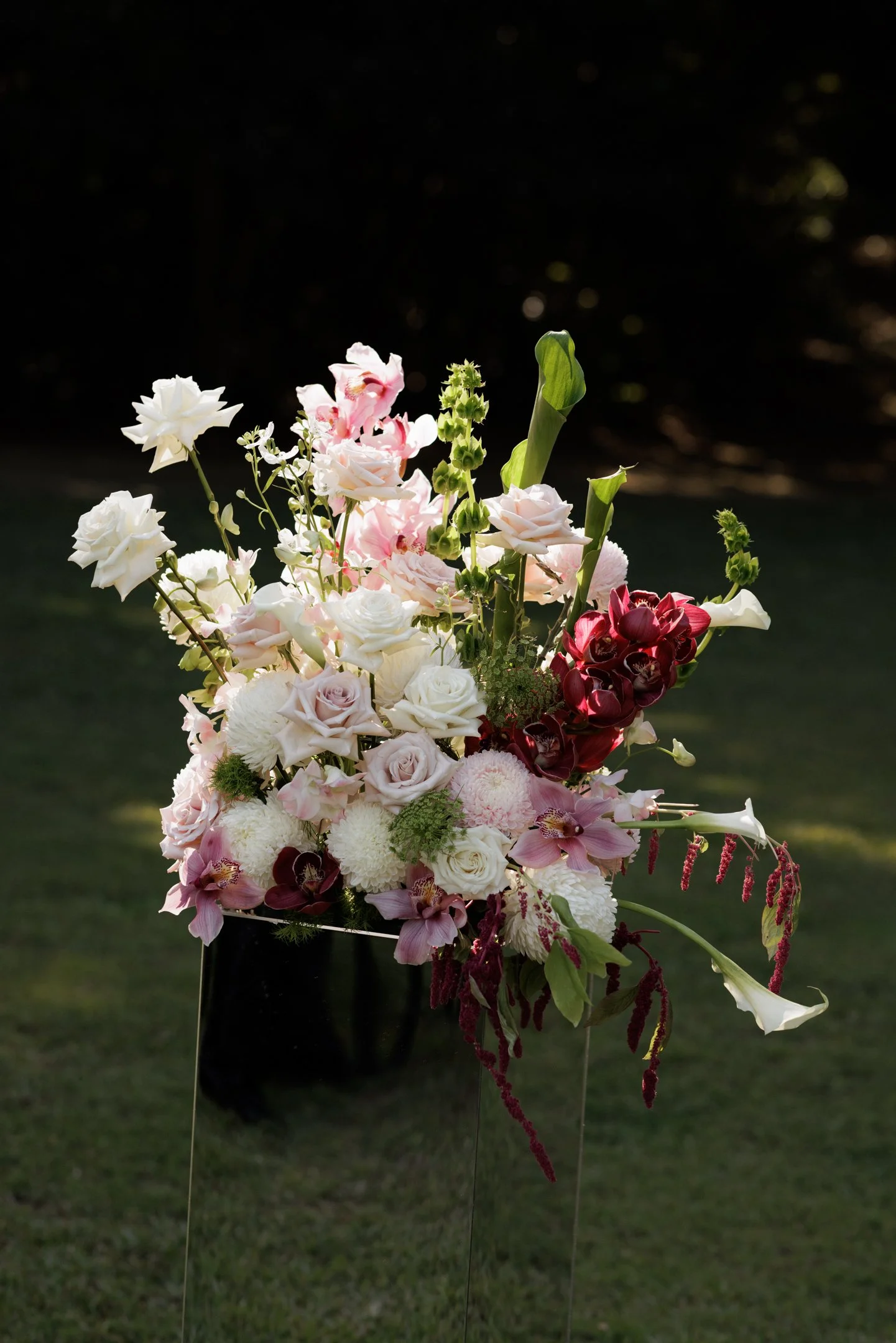 A bouquet of various flowers including roses, calla lilies, and orchids in a black vase set outdoors on grass with a dark background.