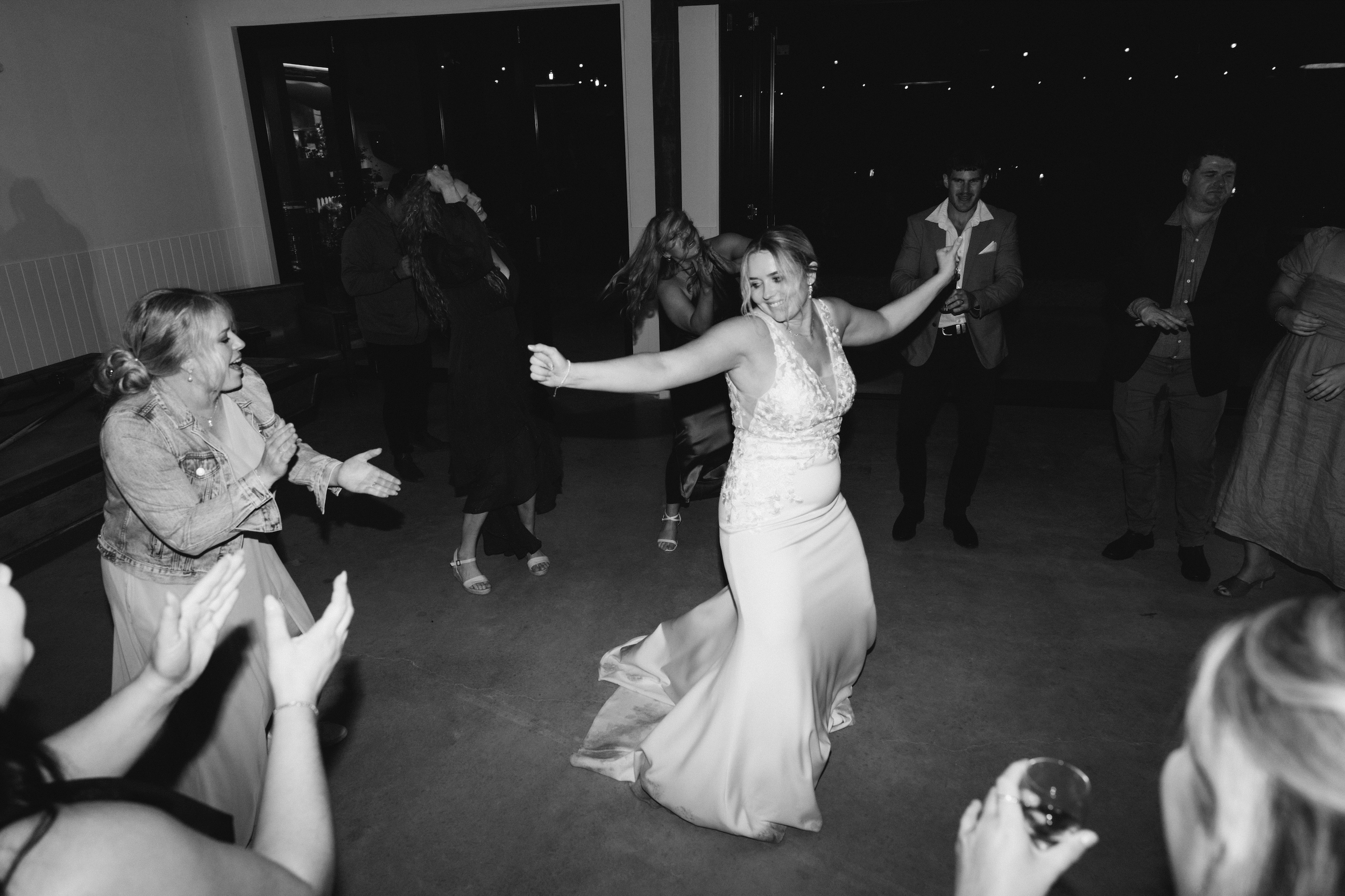 A bride in a wedding dress dancing joyfully with guests at a wedding reception, surrounded by laughing people in a dimly lit venue.