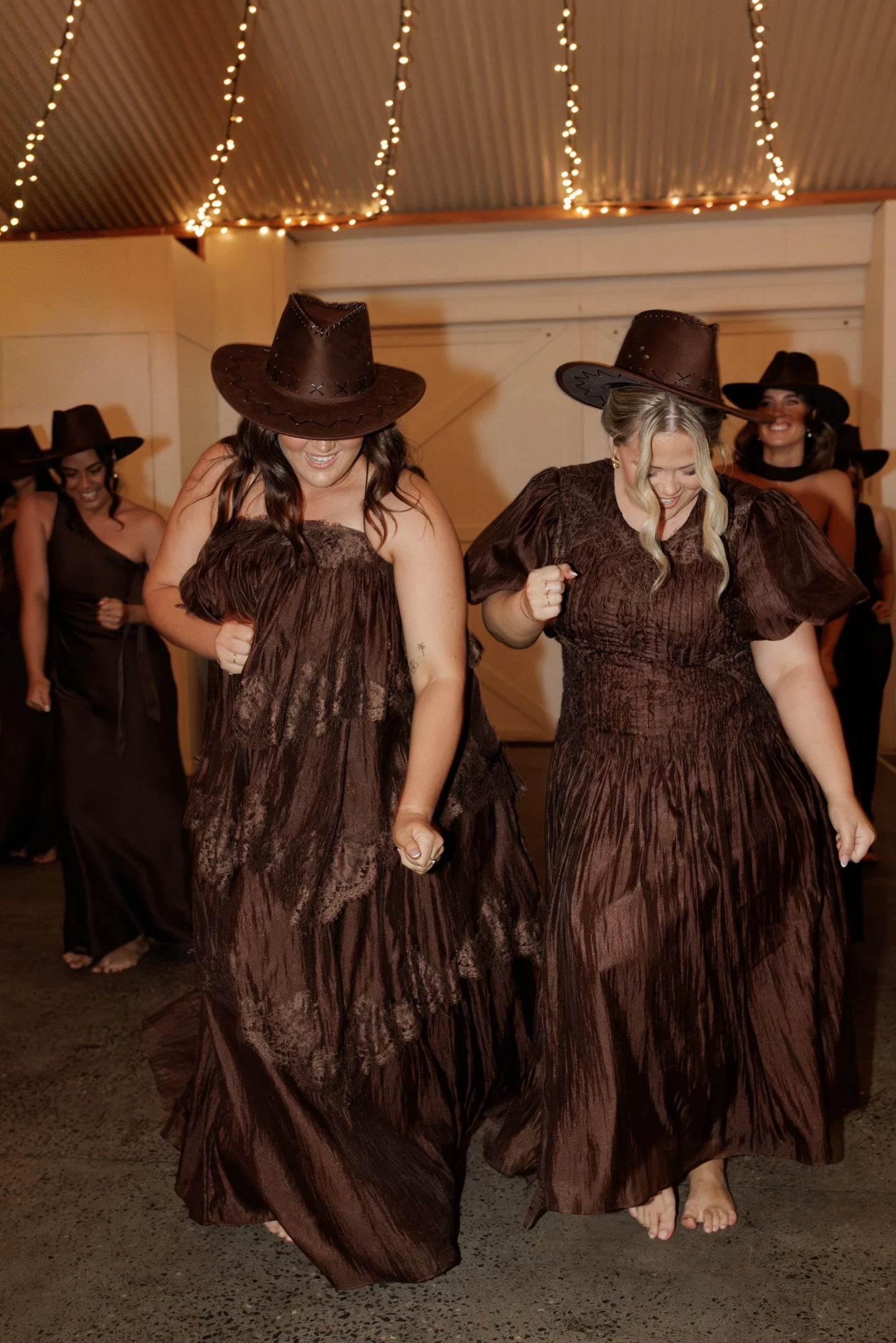 Group of women wearing matching brown dress and cowboy hats, dancing and smiling indoors with string lights overhead.