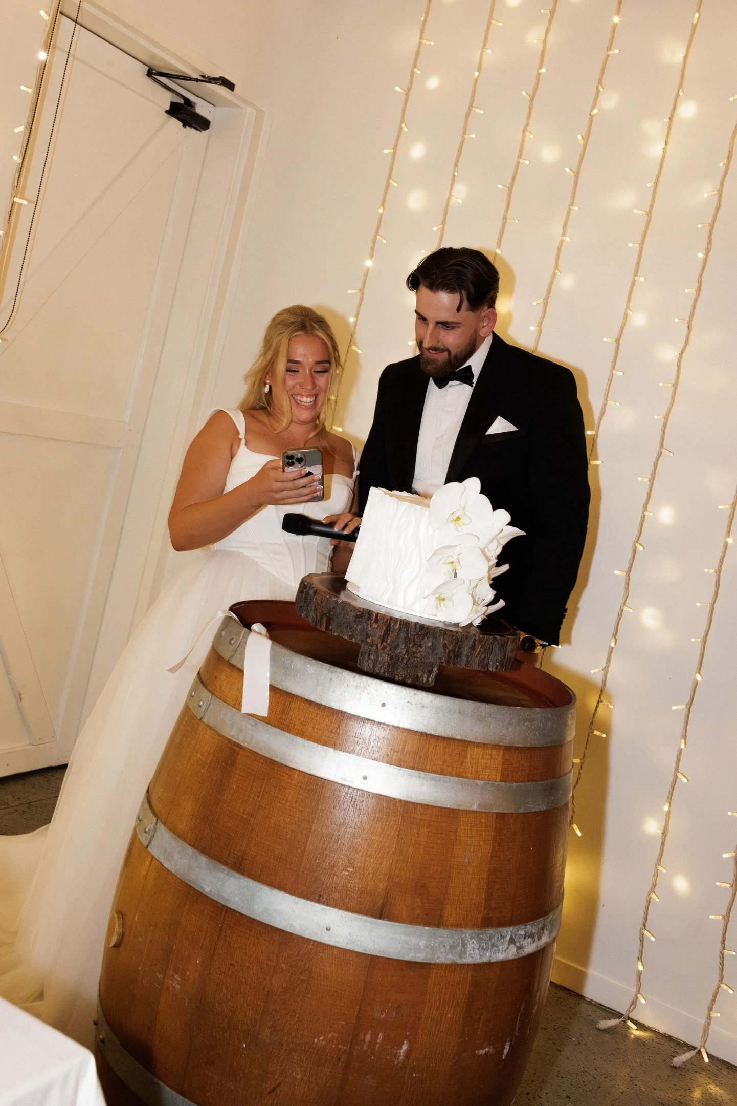 A woman in a white dress and a man in a tuxedo stand behind a large wooden wine barrel that holds a white wedding cake decorated with white orchids. The background features vertical strings of small white lights.